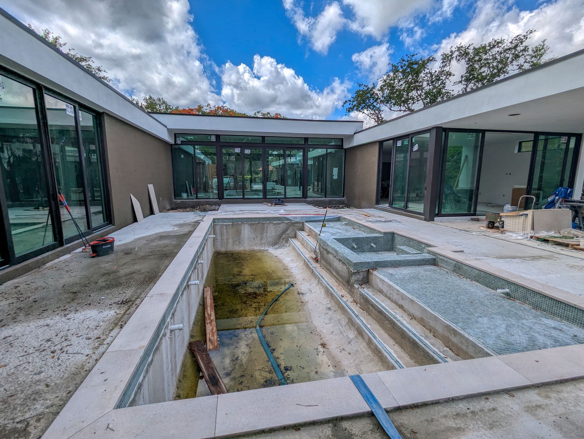 Unfinished pool area with surrounding modern home. Sliding glass doors, blue sky, and construction debris.