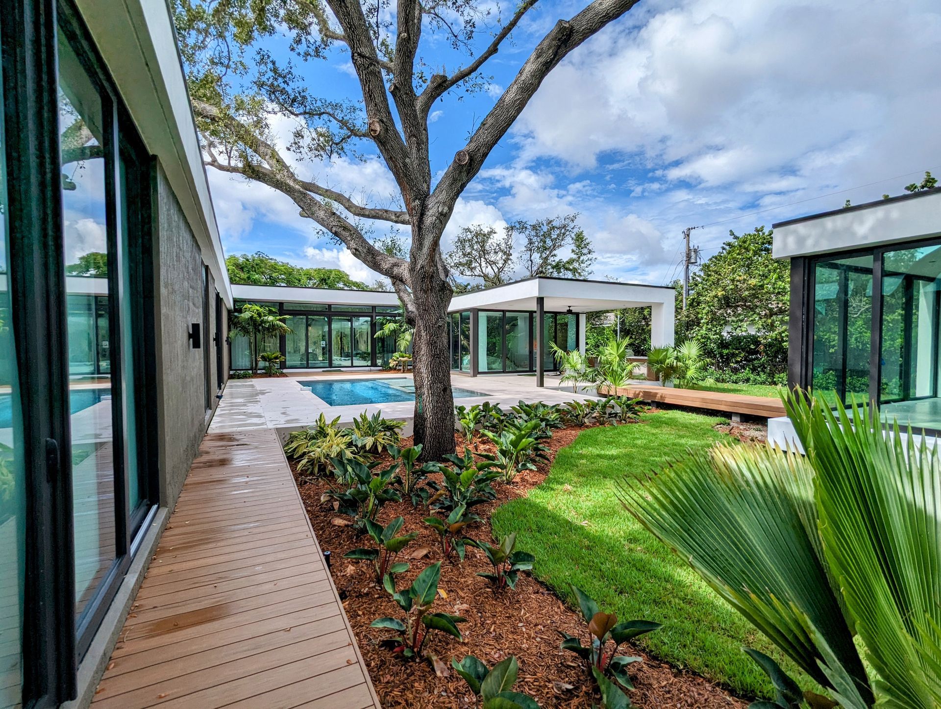 Modern home exterior with pool, large tree, and landscaping under a partly cloudy sky.