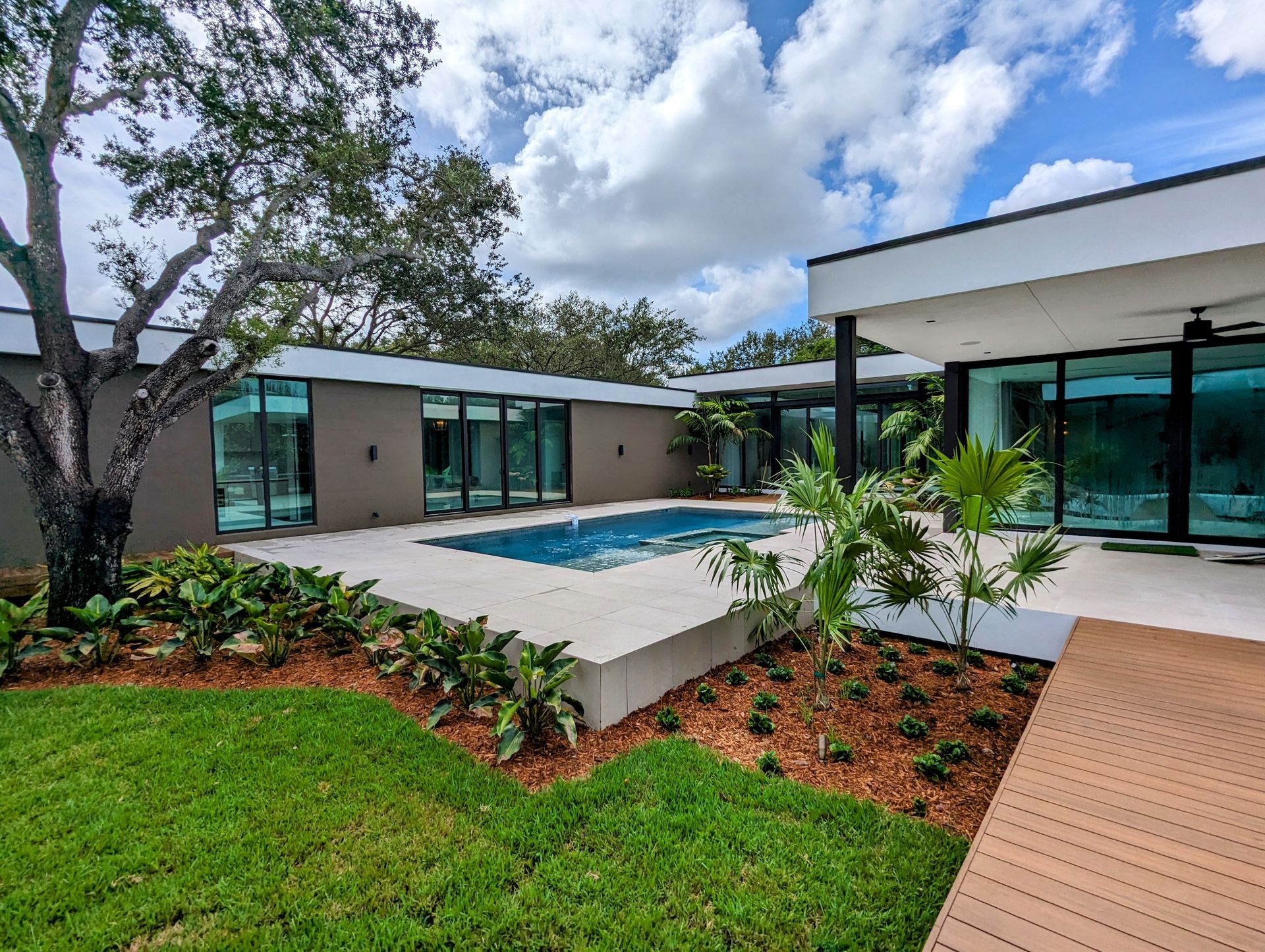 Modern home with pool, glass walls, and lush landscaping on a sunny day.