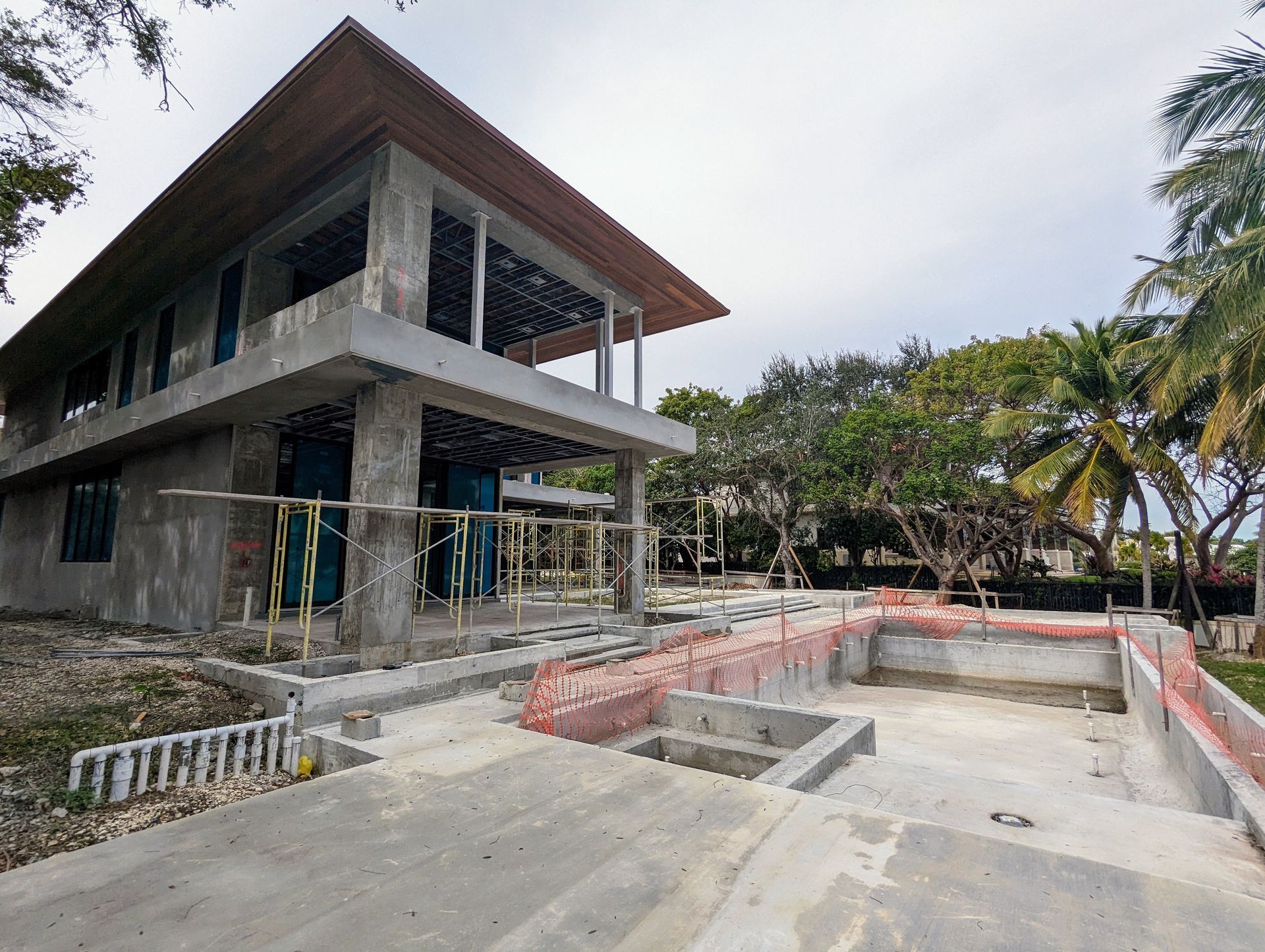 Two-story concrete building under construction with a pool, palm trees, and a cloudy sky.