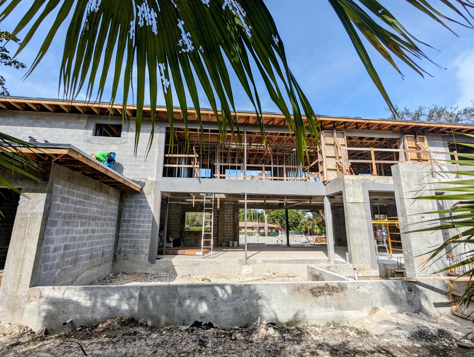 Building under construction, gray concrete, wooden framework, blue sky, tropical foliage in foreground.