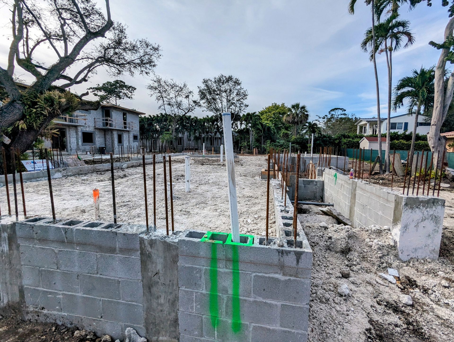 Construction site with concrete block foundation, rebar, and bare ground; residential neighborhood in background.