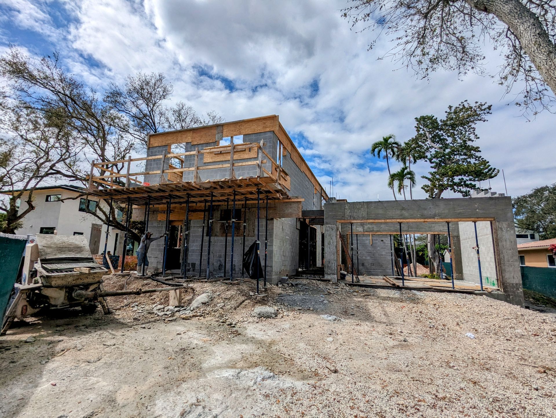 House under construction; concrete structure with scaffolding, exposed wood framing, and debris-covered ground; blue sky backdrop.