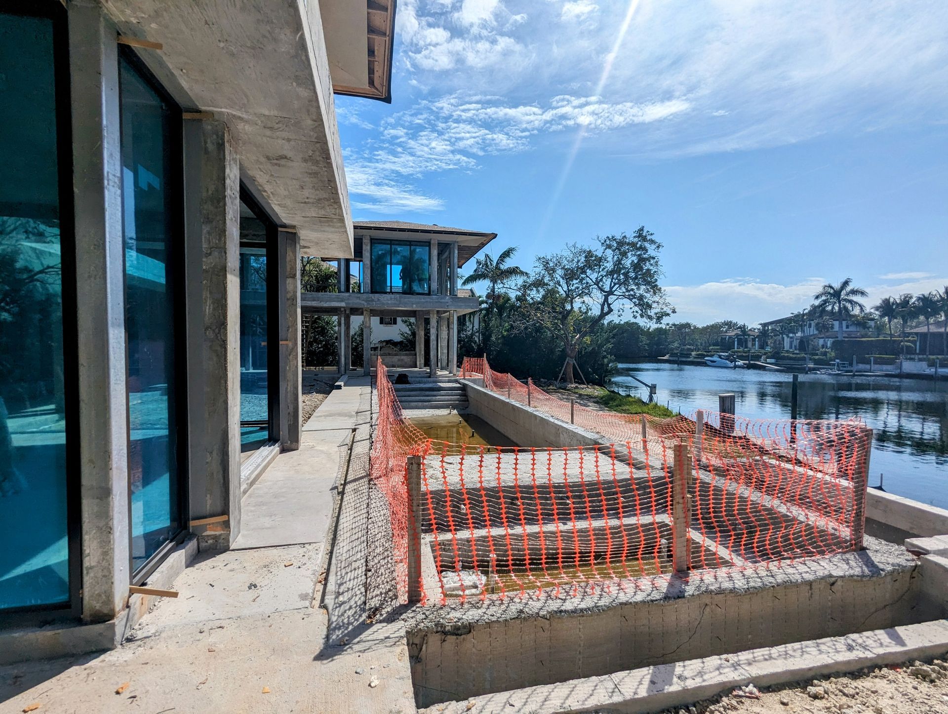 Building under construction, waterfront view. Blue glass windows, orange safety fence, cloudy sky, canal.