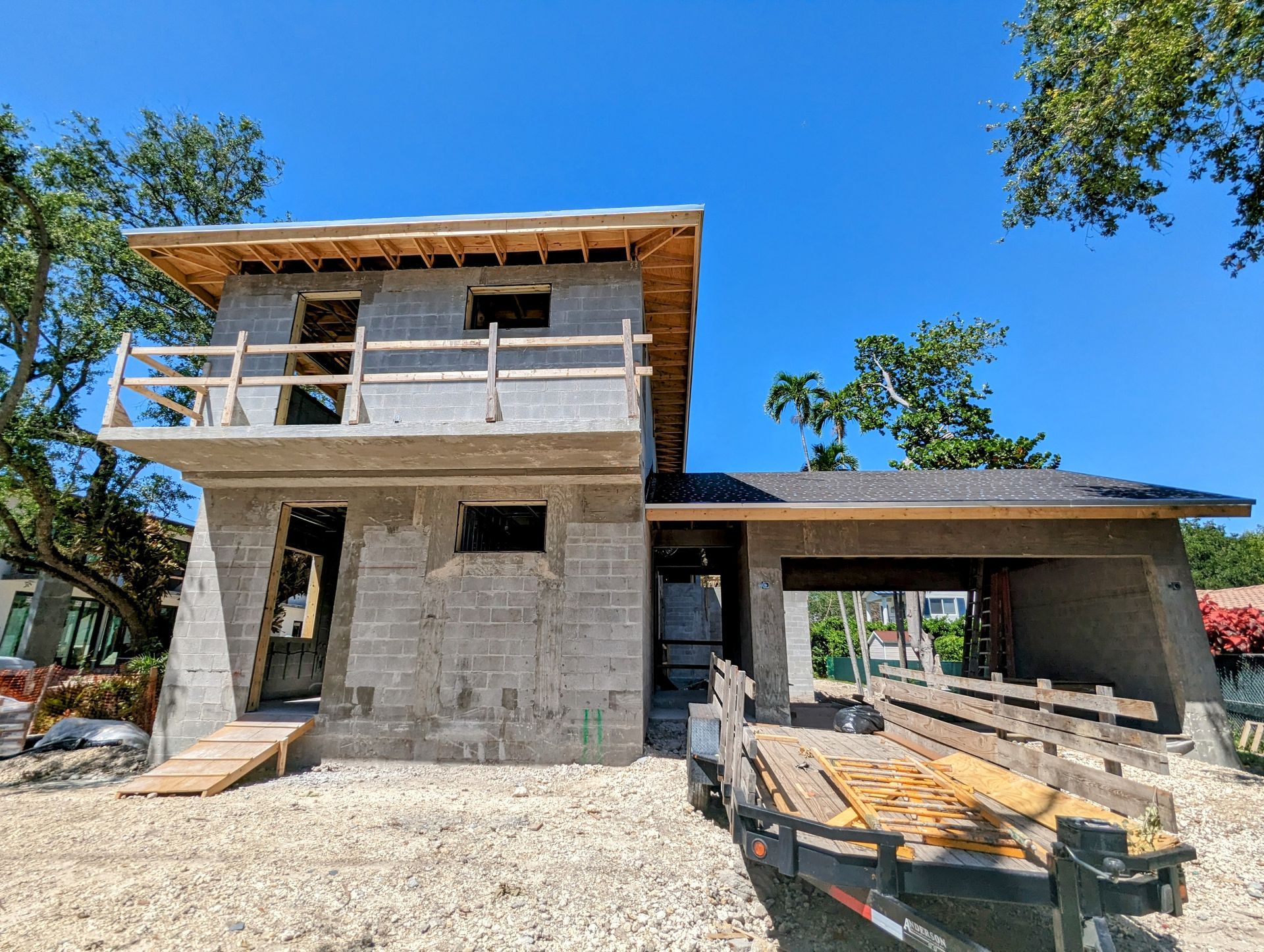 Two-story house under construction, concrete block walls, wooden roof frame. Trailer with lumber in front. Sunny day.