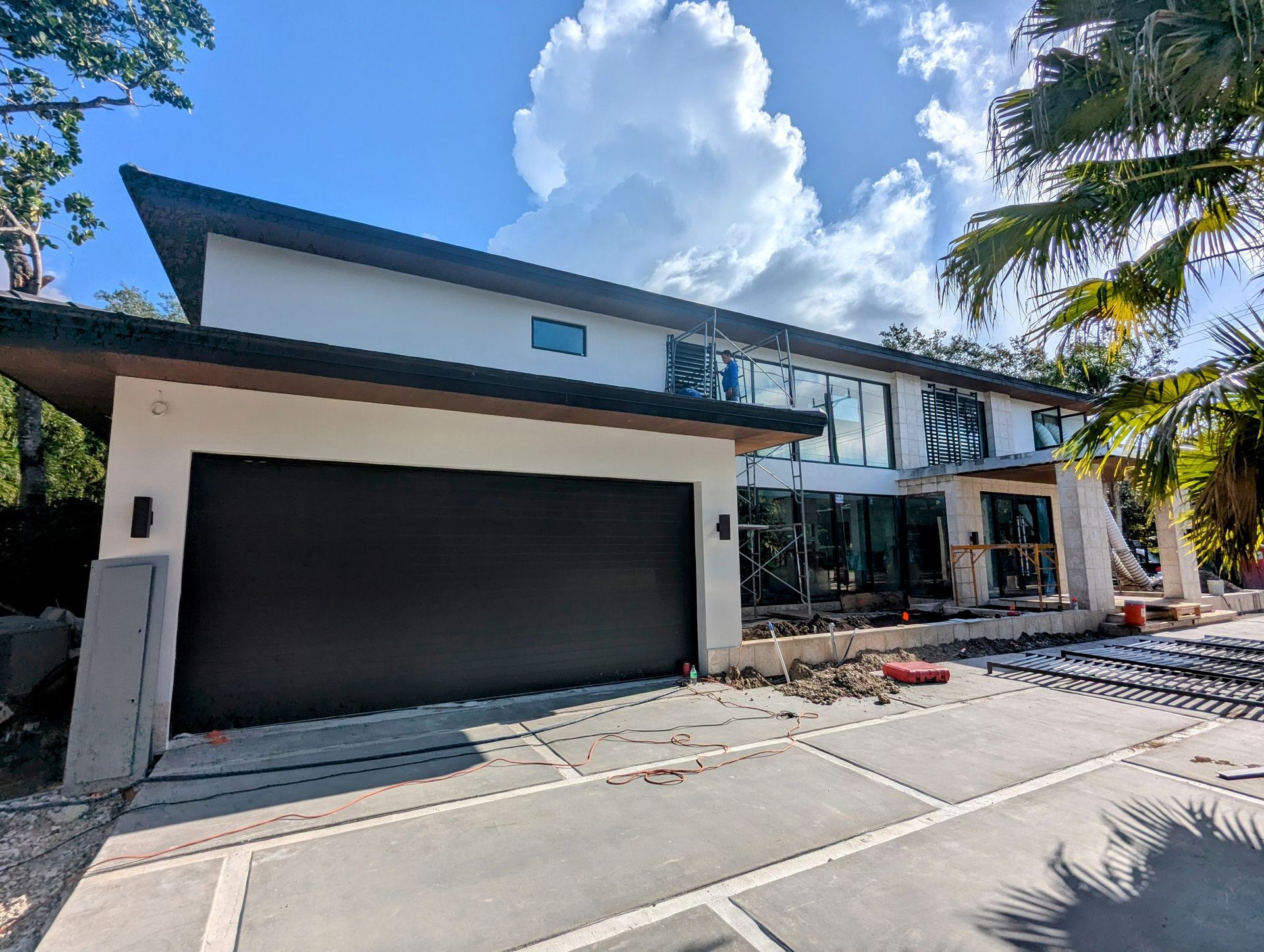 Modern two-story house under construction. Brown garage door, white walls, large windows. Bright sky, palm trees.