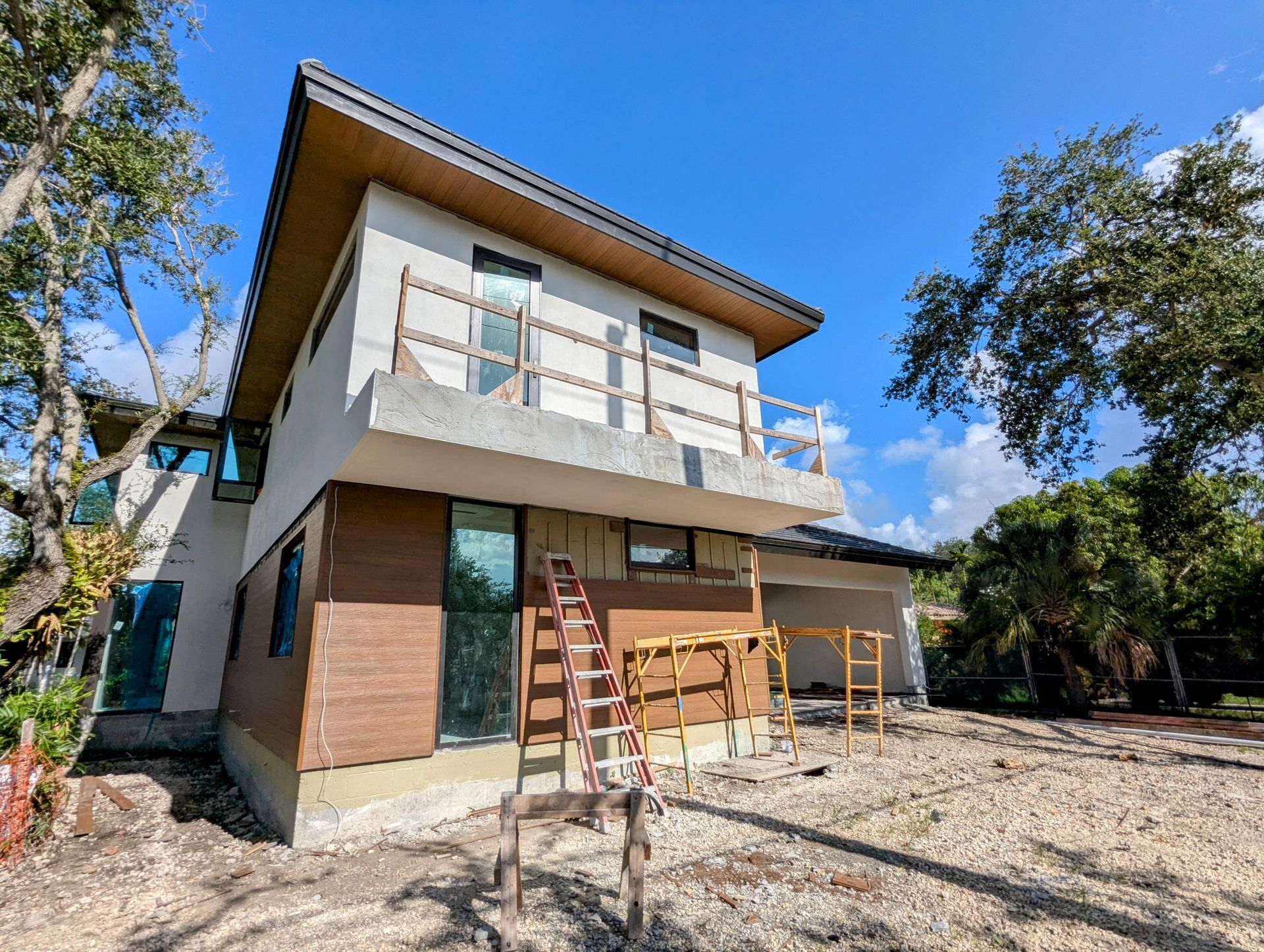 Two-story house under construction. Light-colored stucco and brown siding, unfinished balcony, ladder in front, bright blue sky.