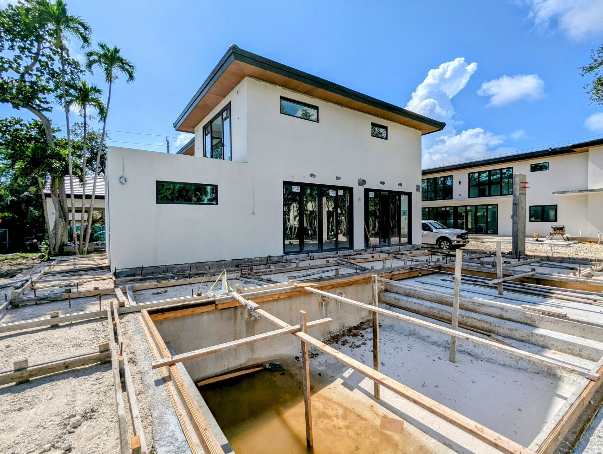 Two-story white house under construction, with a concrete foundation in the foreground and blue sky.