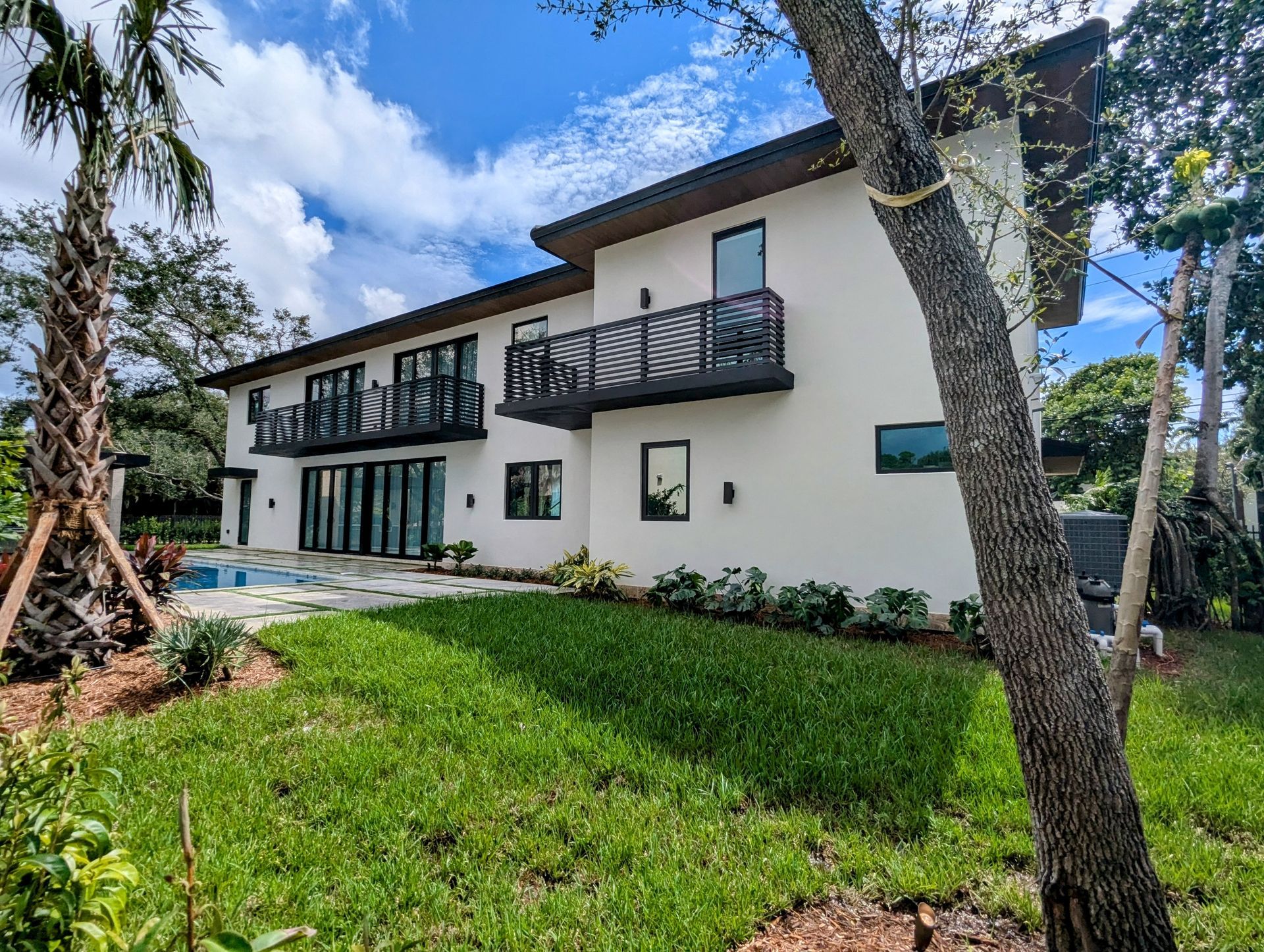 Modern white house with dark trim and balconies, surrounded by greenery and a pool.