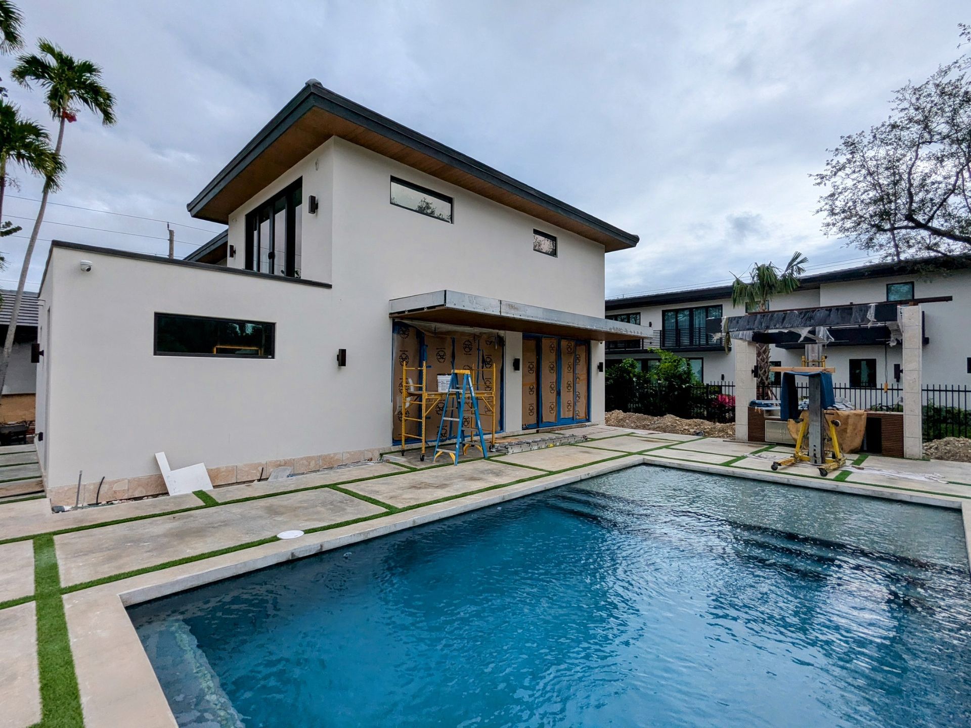 Modern white house with a pool. Dark roof, wooden doors, and green grass accents. Overcast sky.