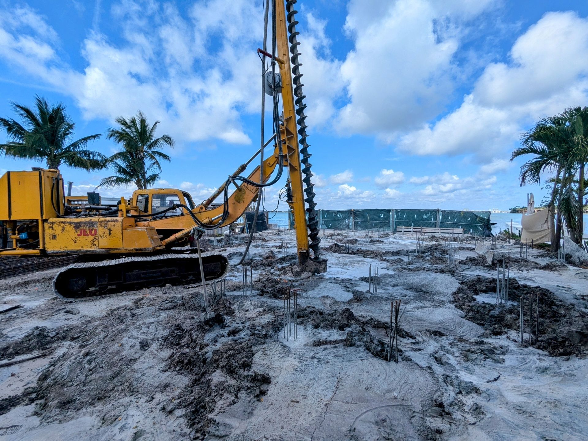 Yellow construction drill on a sandy worksite by the water under a cloudy sky.