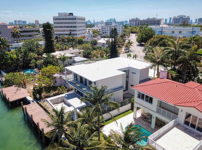 Aerial view of waterfront homes with docks, palm trees, and city skyline in the background.