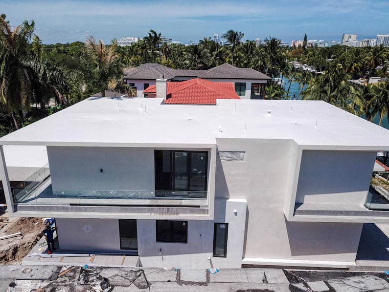 Modern white house with a flat roof under construction; a worker stands below.