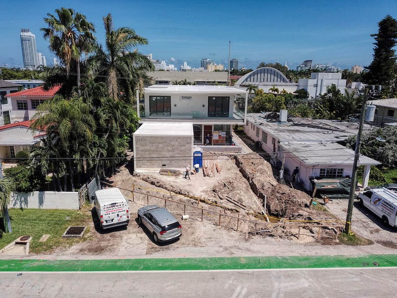 Two-story modern house under construction. Miami, Florida. White top floor, stone-colored ground floor, surrounded by dirt and vehicles.