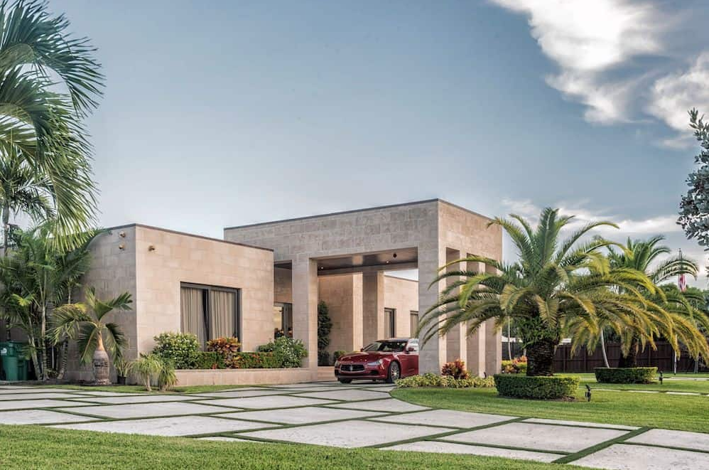 Modern beige house with car parked beneath a covered entryway. Lush greenery, palm trees, blue sky.