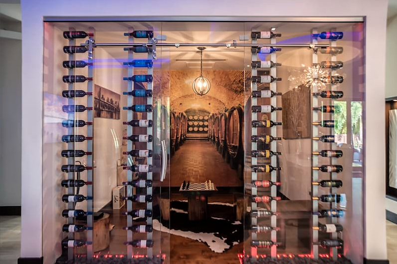 Glass-enclosed wine cellar with rows of bottles on metal racks, a long tunnel-like interior, and colorful lighting.