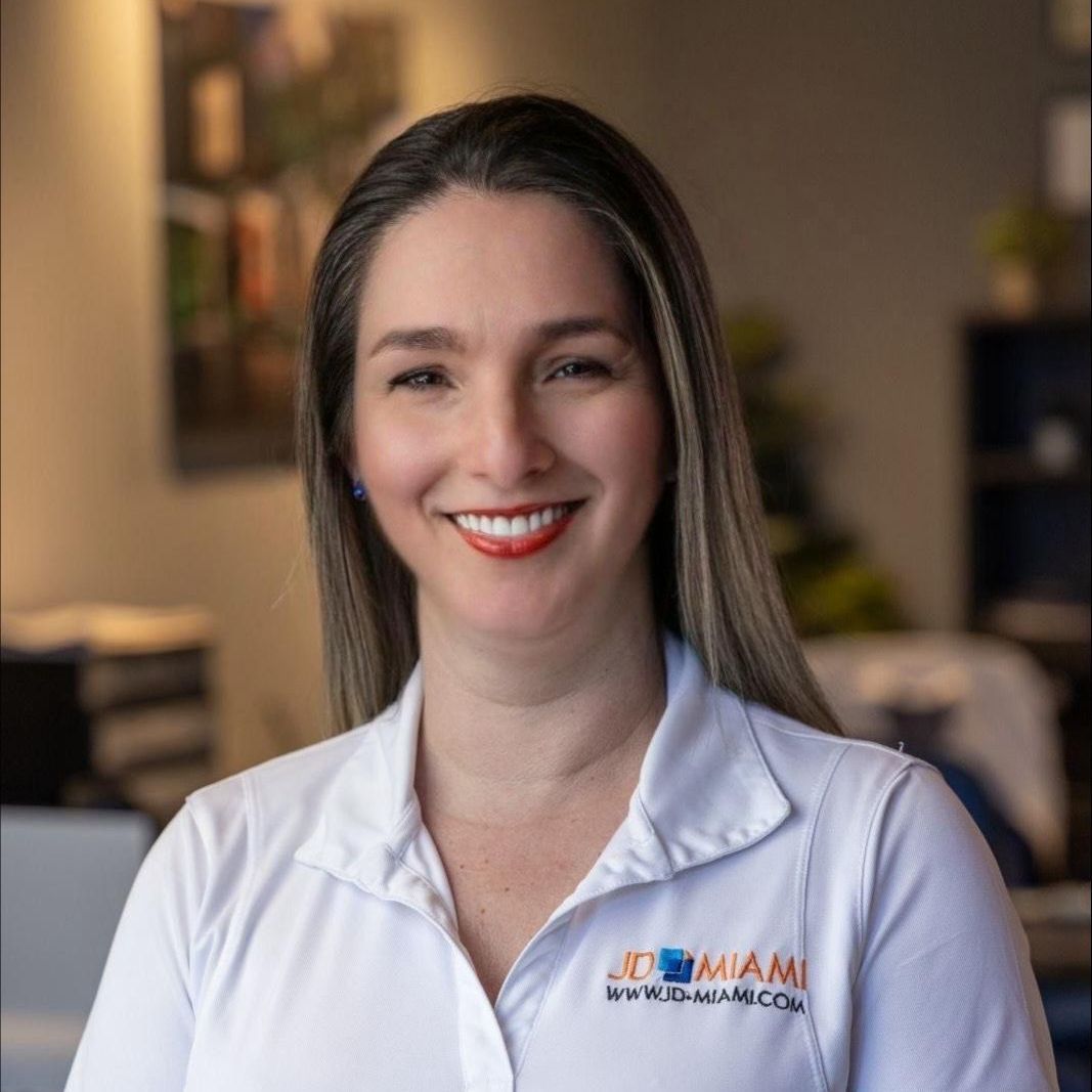 Woman smiling, wearing a white shirt with a logo, in an office setting.