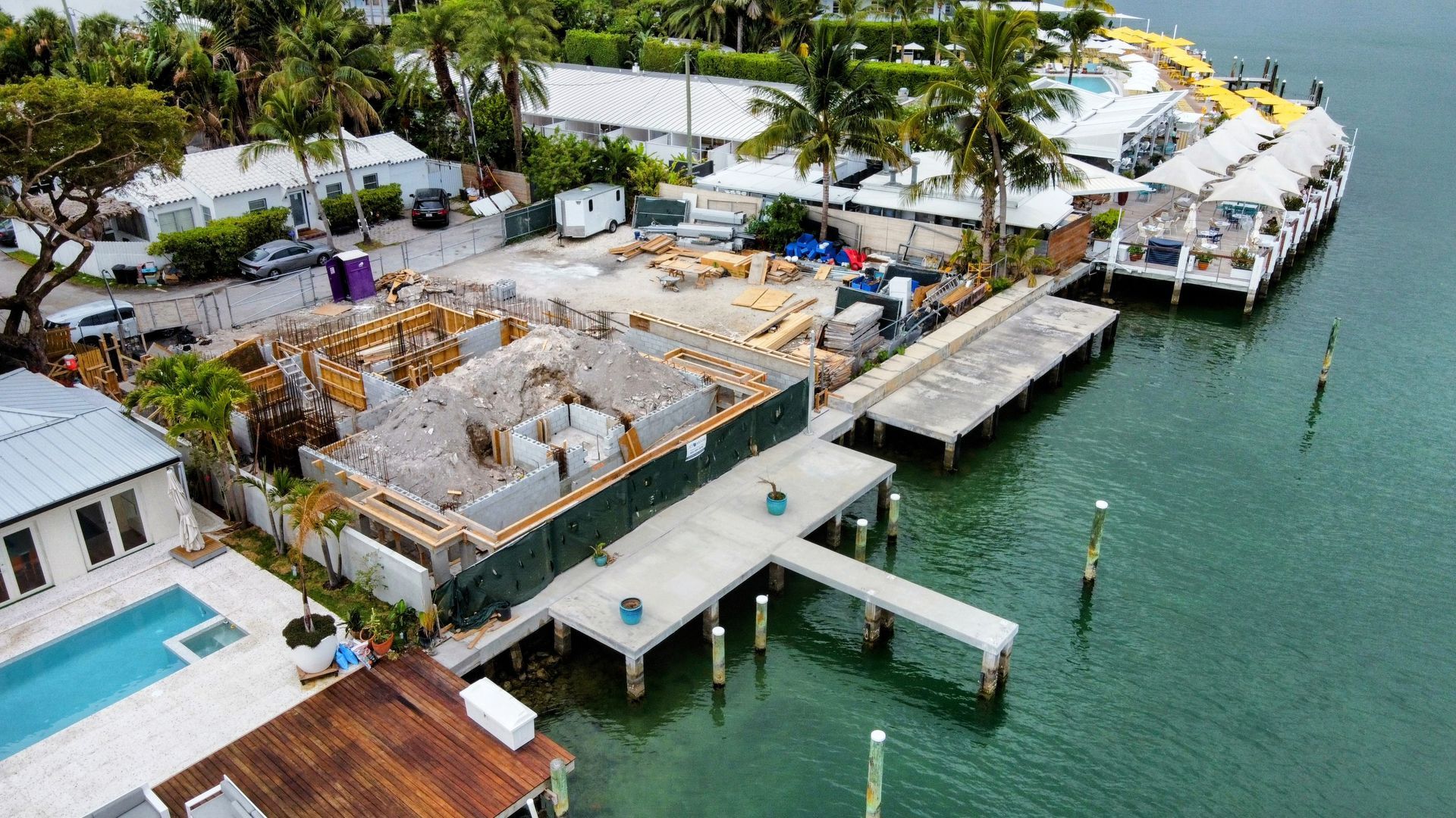 Construction site on waterfront, with unfinished building, dock, boats, and partial view of a pool.