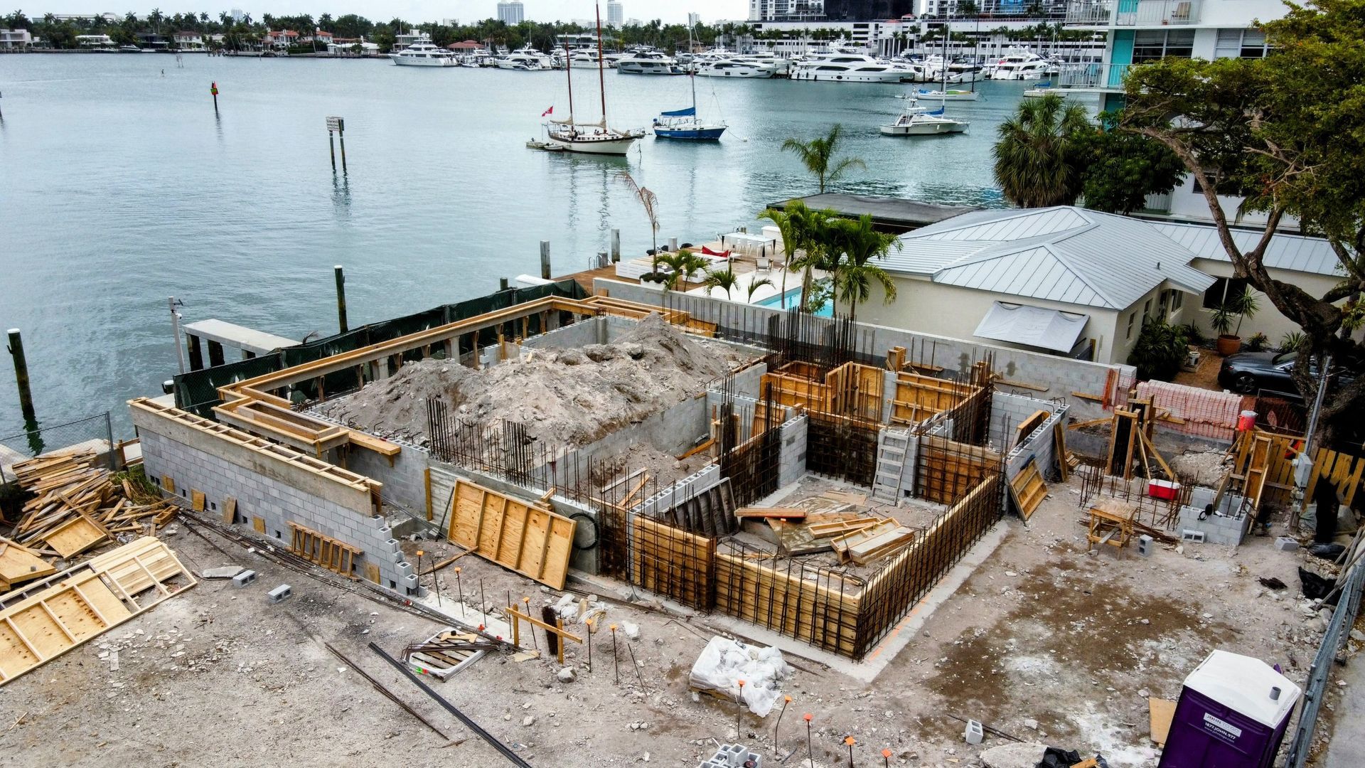 Construction site on waterfront, forming building foundations with wooden frames, boats in background.