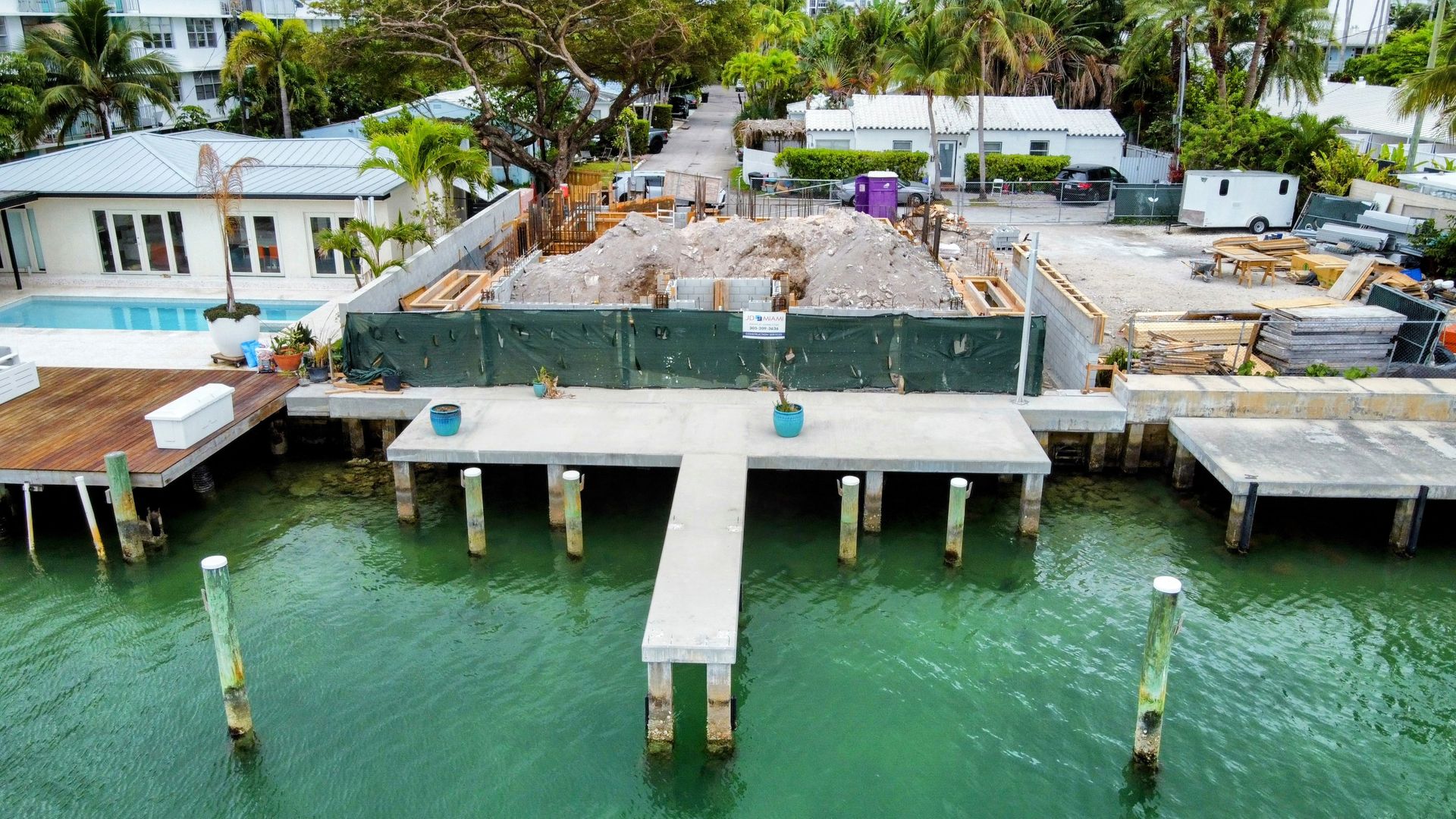 Construction site over water; concrete dock, fence, debris, houses, and palm trees.