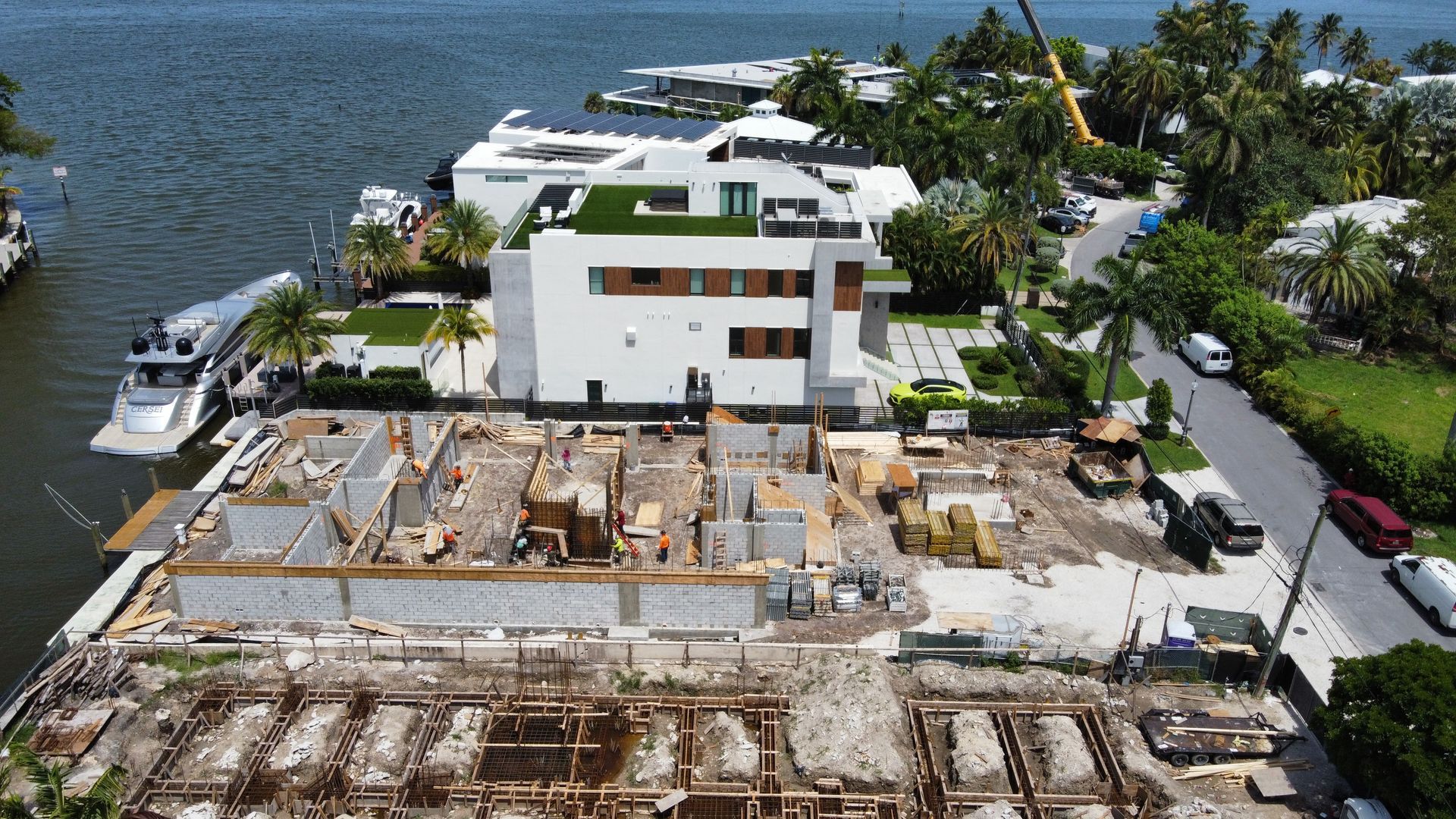 Construction site next to a white house with green roof and boats in water.