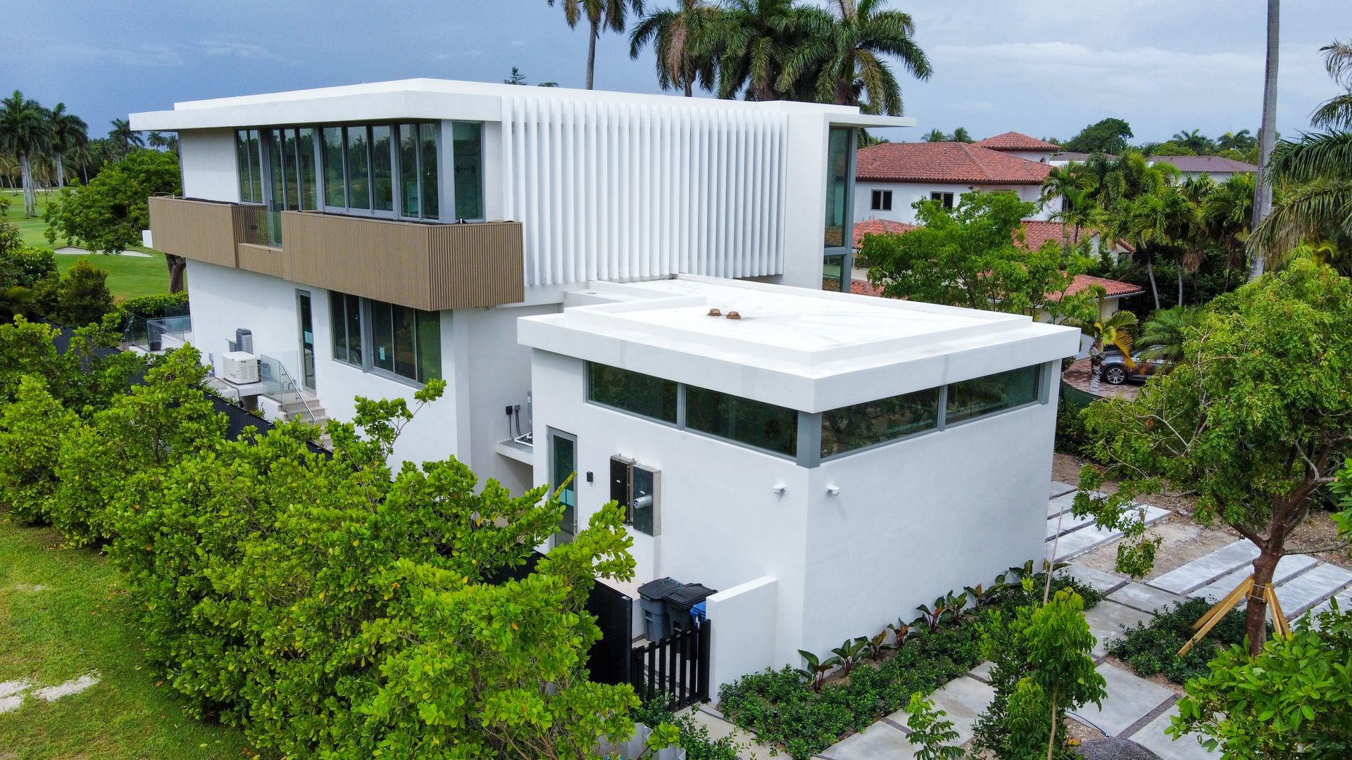 Modern white house with flat roof, glass windows, and vertical siding, surrounded by trees and greenery.
