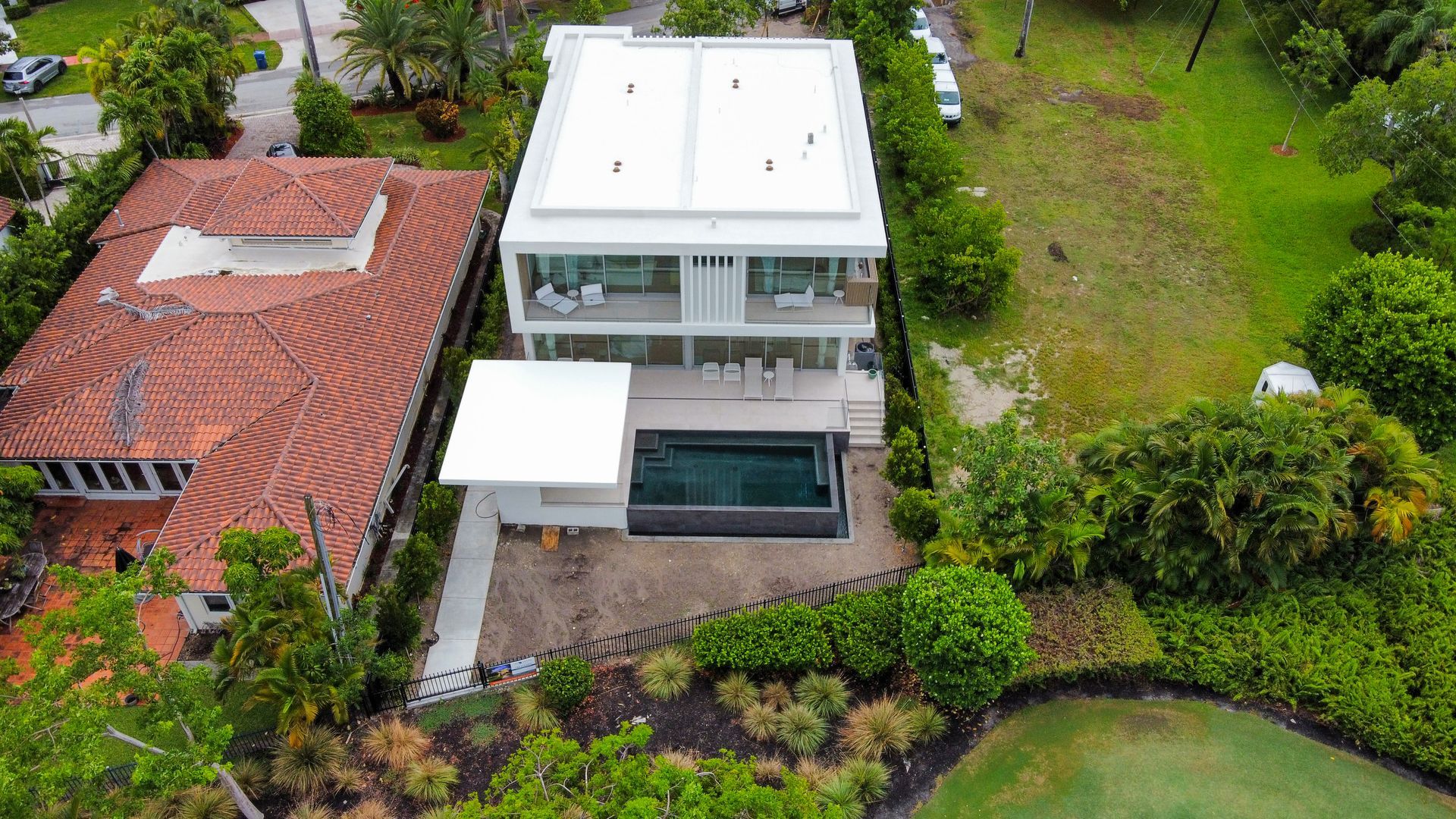 Modern white house with a pool, next to a house with an orange roof, surrounded by greenery.