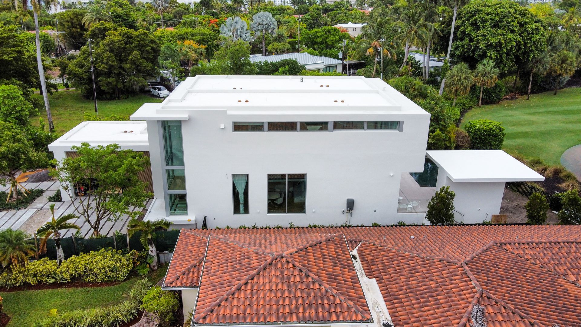 White modern home with flat roof, surrounded by green trees and a red-tiled roof in the foreground.