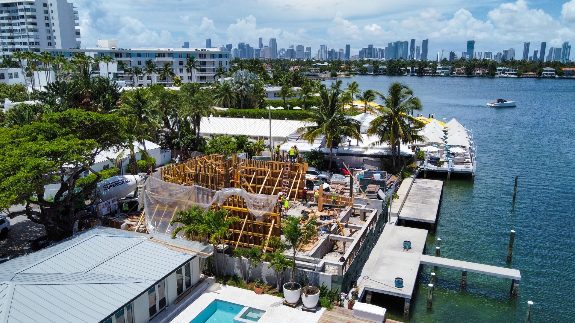 Aerial view of a coastal construction site with stacked lumber, docks, and a city skyline in the background.