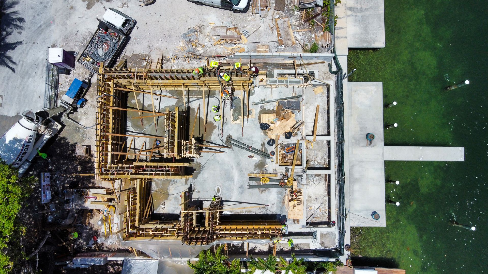 Overhead view of a house under construction by water. Concrete foundation and wood framing are visible.