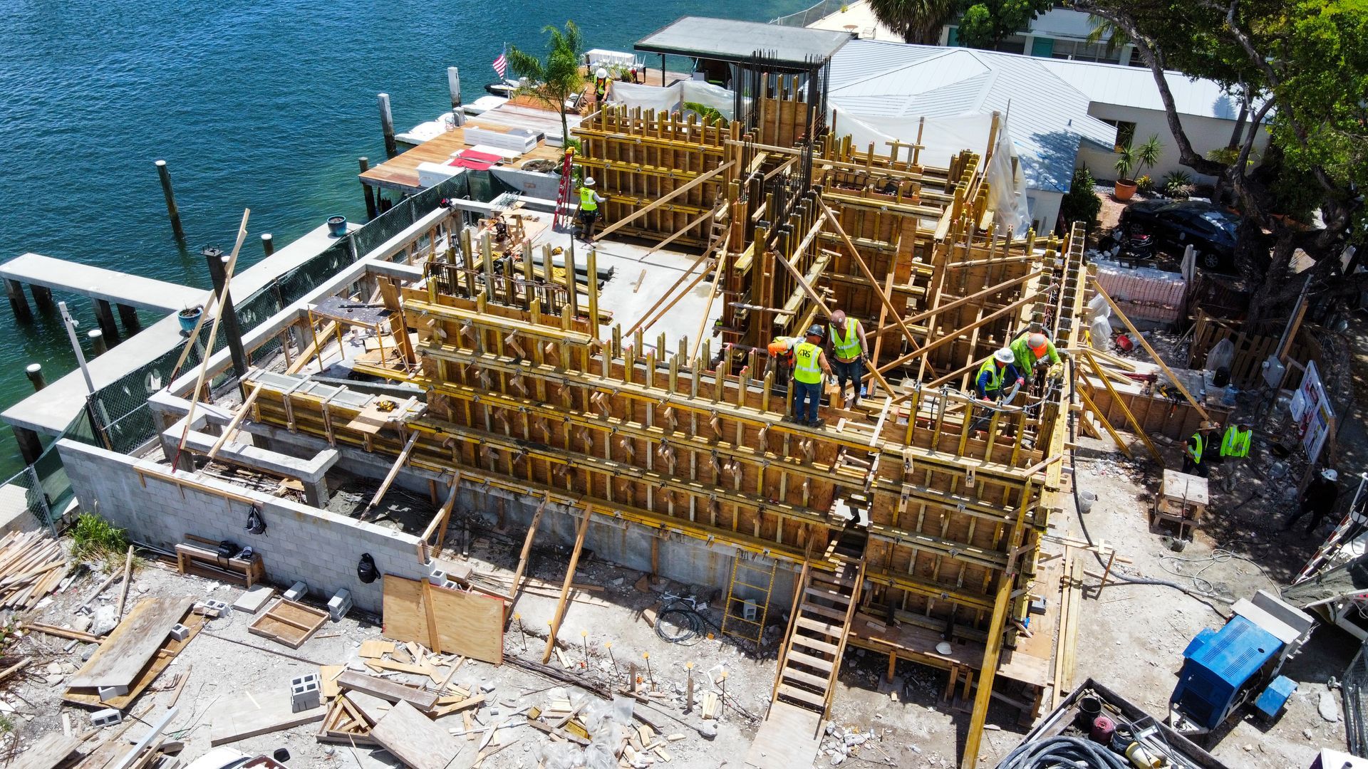 Construction site by water: workers, wooden forms, concrete base, bright day.