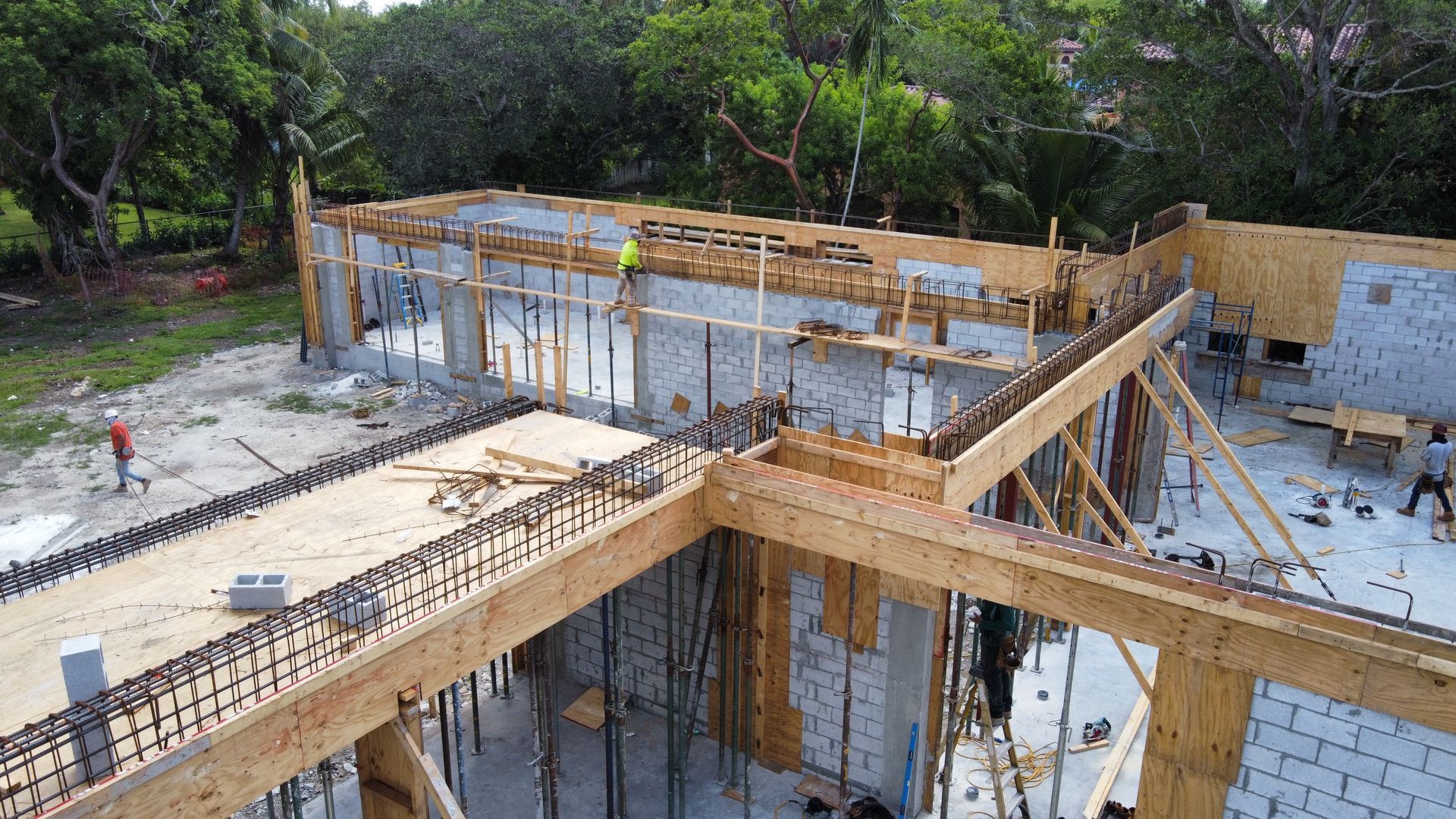 Construction site with wooden forms and rebar for concrete walls, workers present, green trees in background.