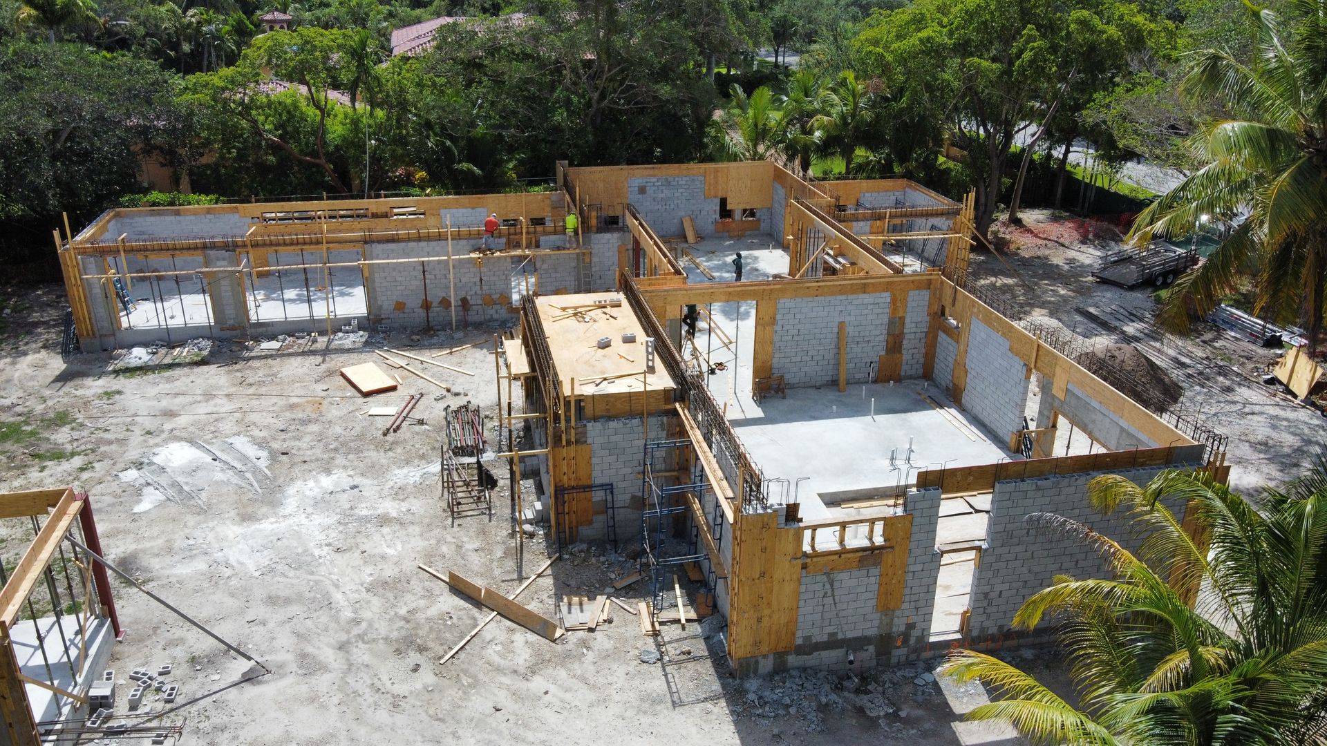 Construction site: wooden frames and concrete walls of a house, surrounded by dirt and trees.
