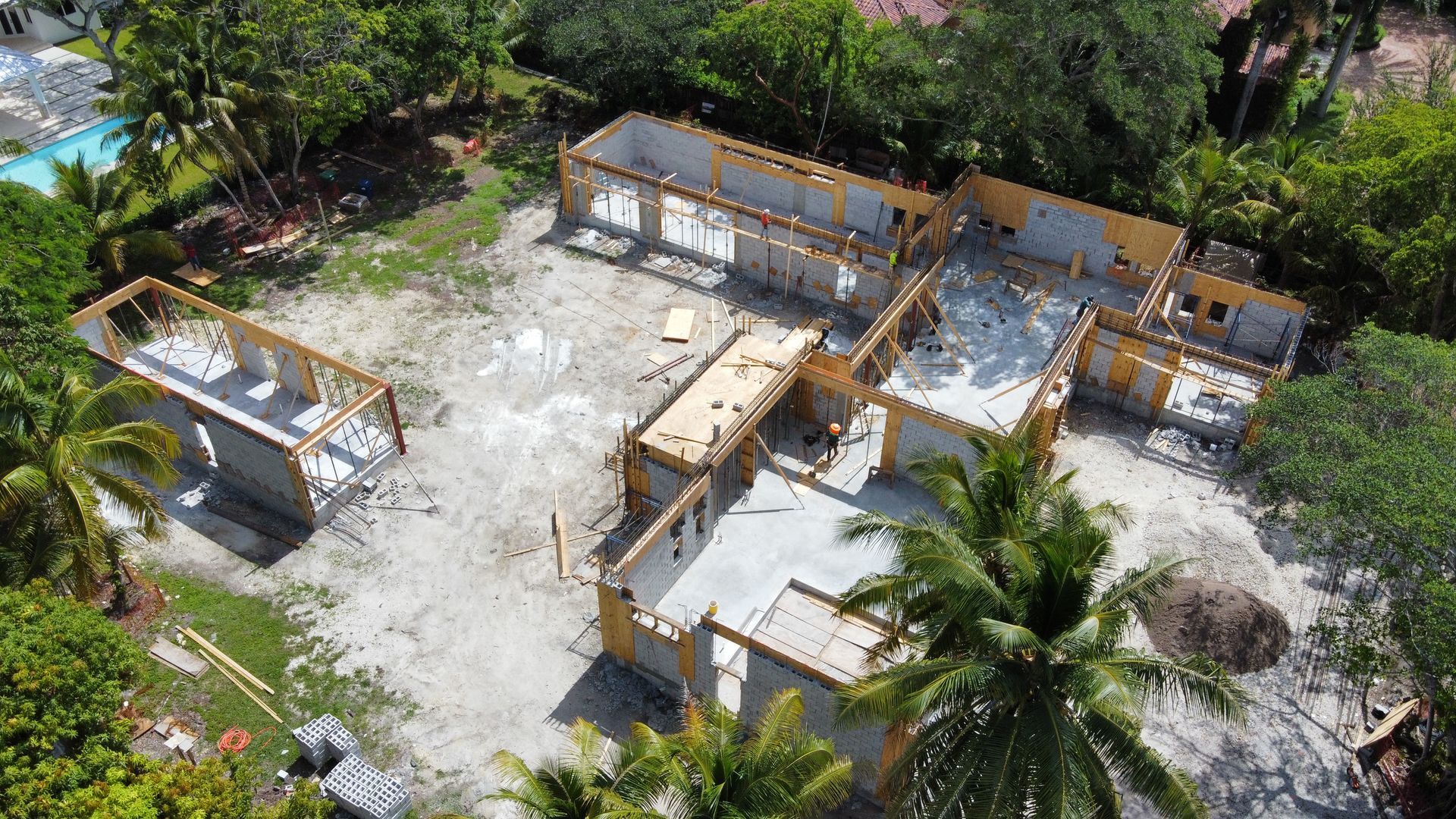 Aerial view of a house under construction with wood framing and concrete foundation, surrounded by trees.