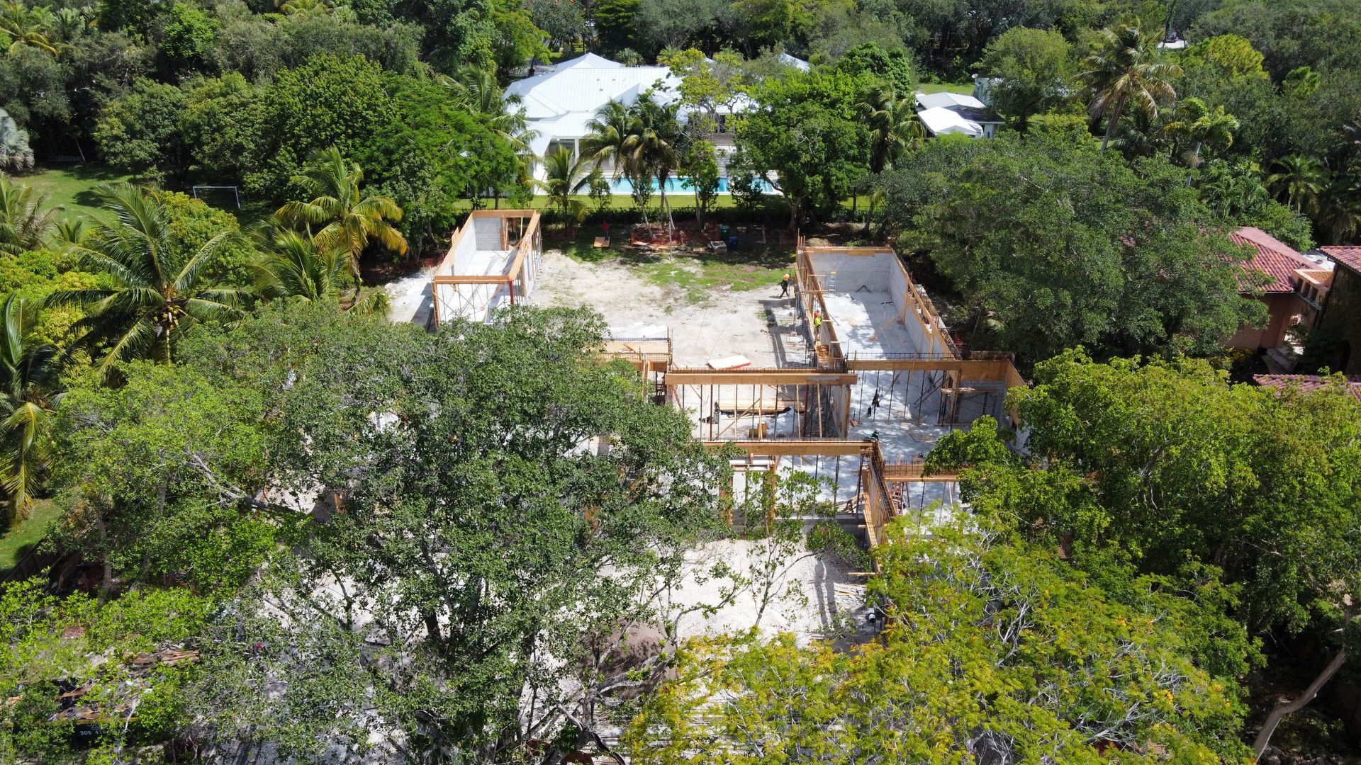 Aerial view of a home under construction surrounded by lush green trees, with existing homes visible in the background.