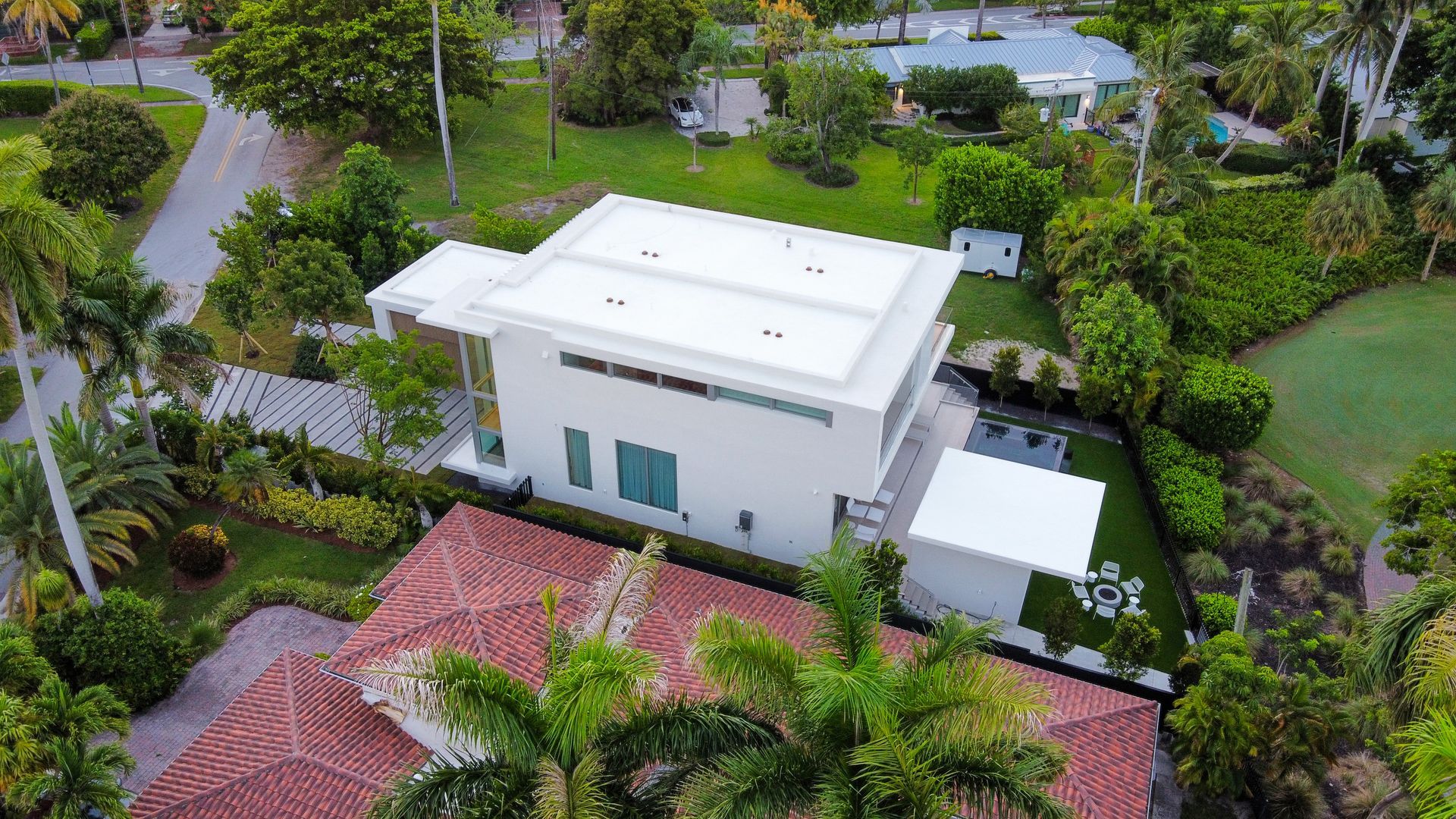 Aerial view of a modern white house with a flat roof, surrounded by green foliage and neighboring homes.