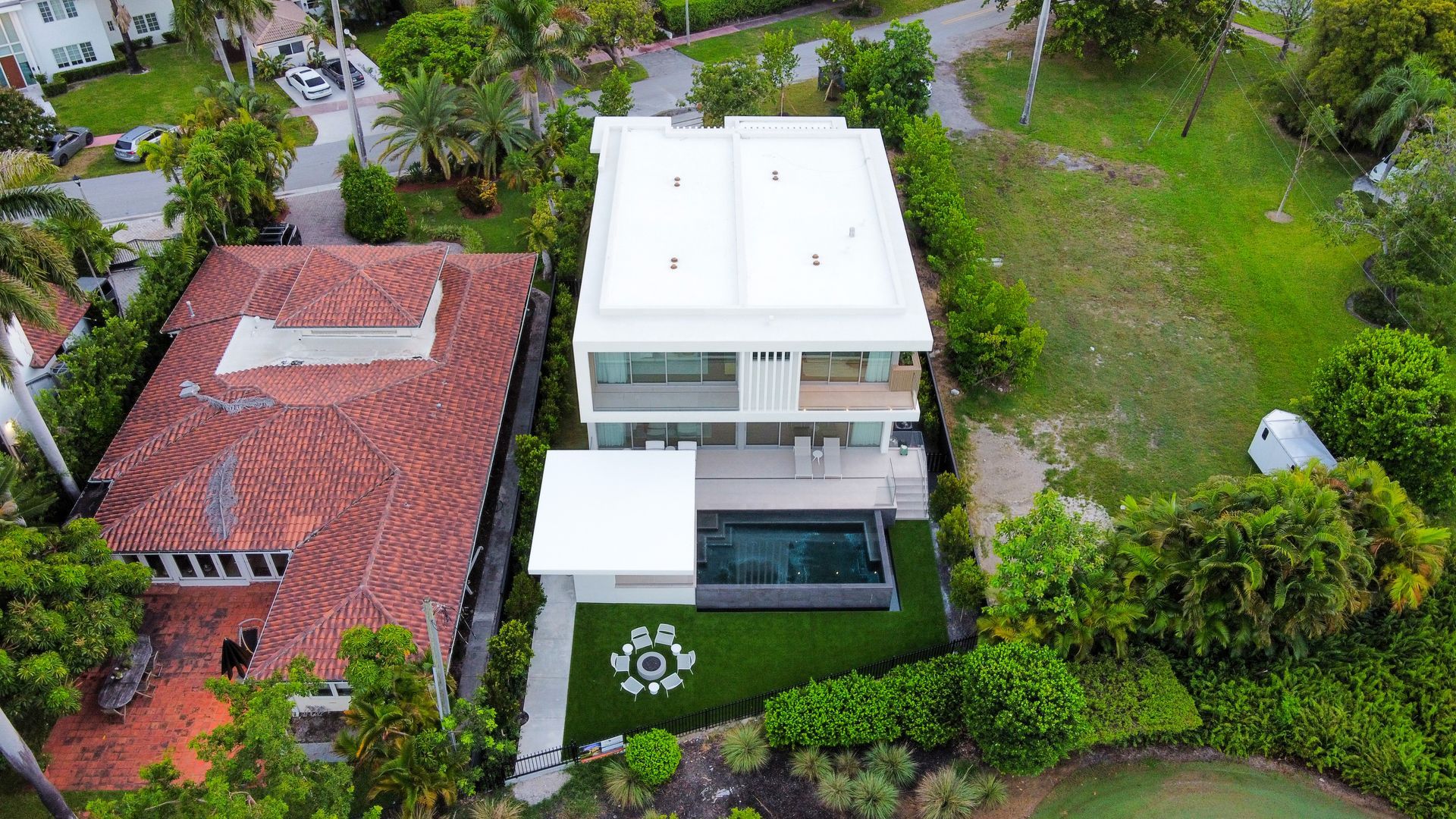 Aerial view of two homes; one with red tiled roof, other modern white with pool.