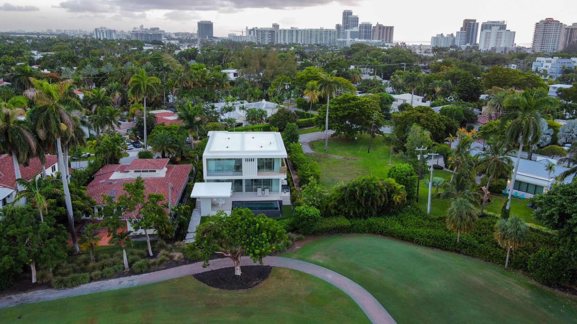 Aerial view of a modern white house surrounded by lush greenery and other homes, with a cityscape in the background.
