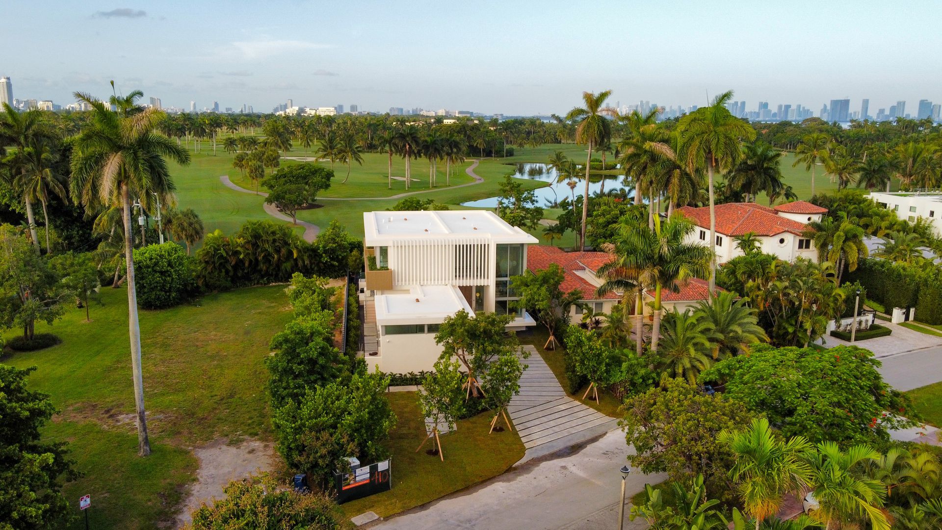 Modern white house with glass windows and a palm tree-lined golf course in the background. City skyline visible.