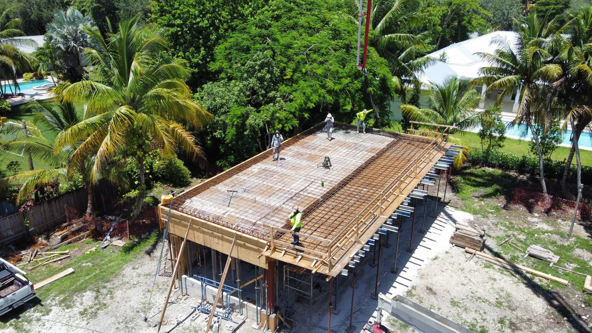 Construction workers on a wooden concrete form, surrounded by green trees and structures, near a pool.