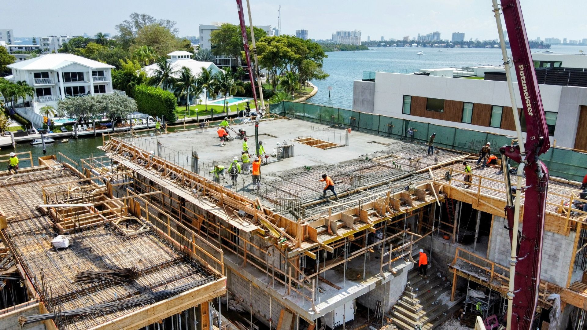 Construction site: Workers pouring concrete onto a building's roof with a pump, waterfront in background.