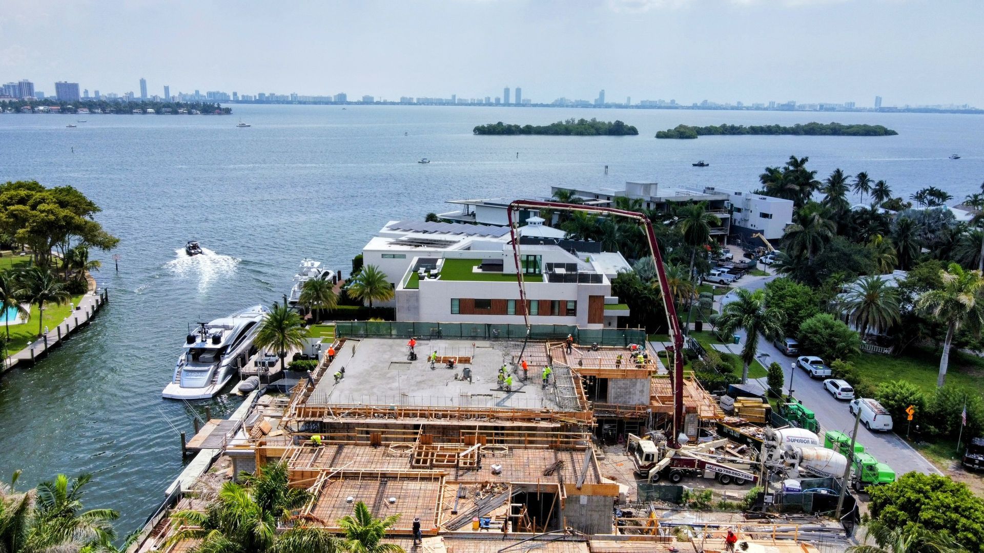 Construction site of a waterfront home with workers, crane, and materials; water and city skyline in the background.