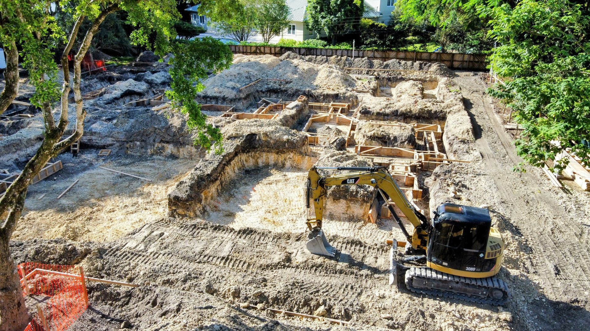 Excavator in a construction site digging a foundation; dirt, stone, and trees visible.