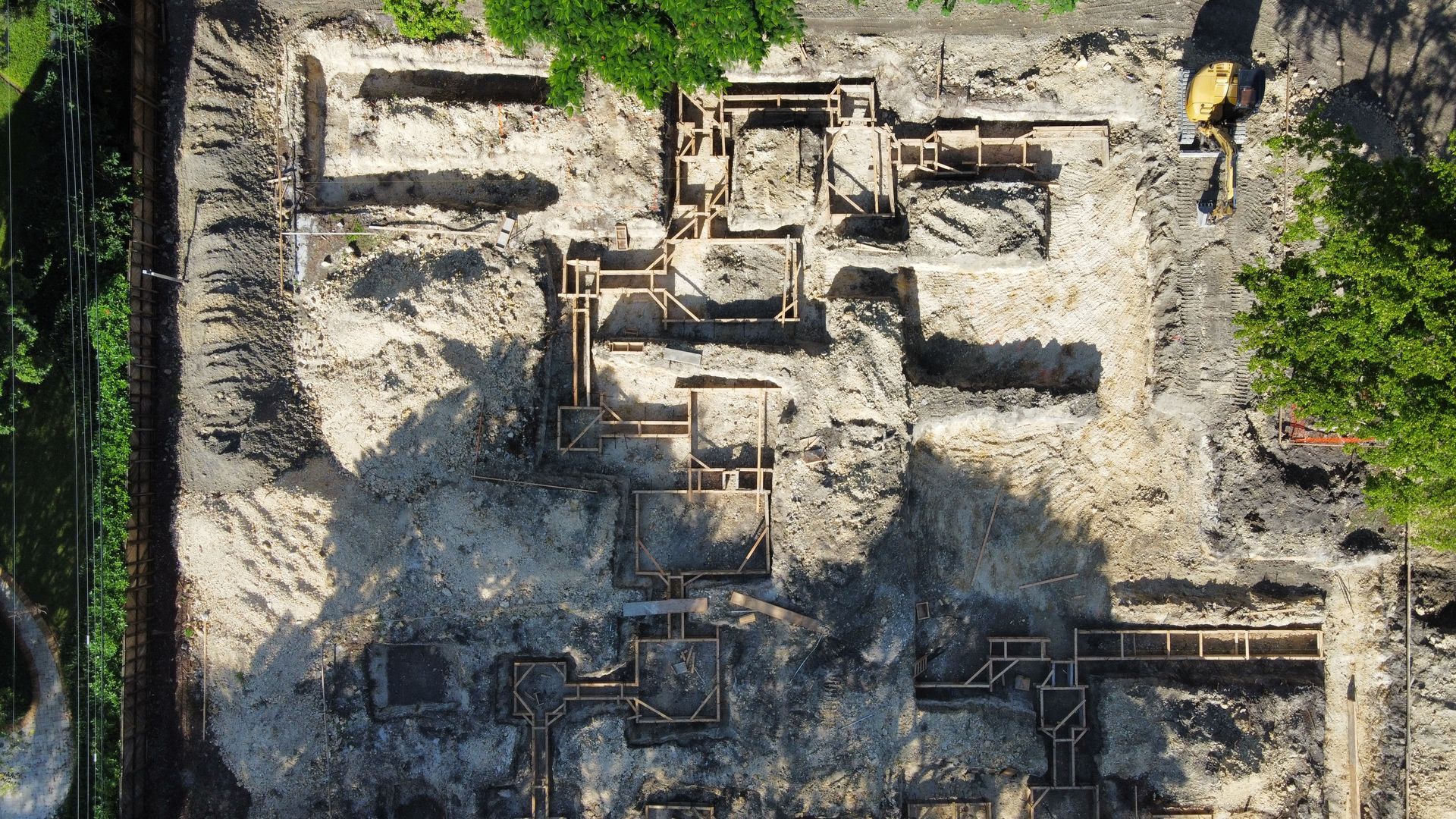 Overhead view of a construction site with excavated foundation outlines, partially filled with soil; trees and excavator are also visible.