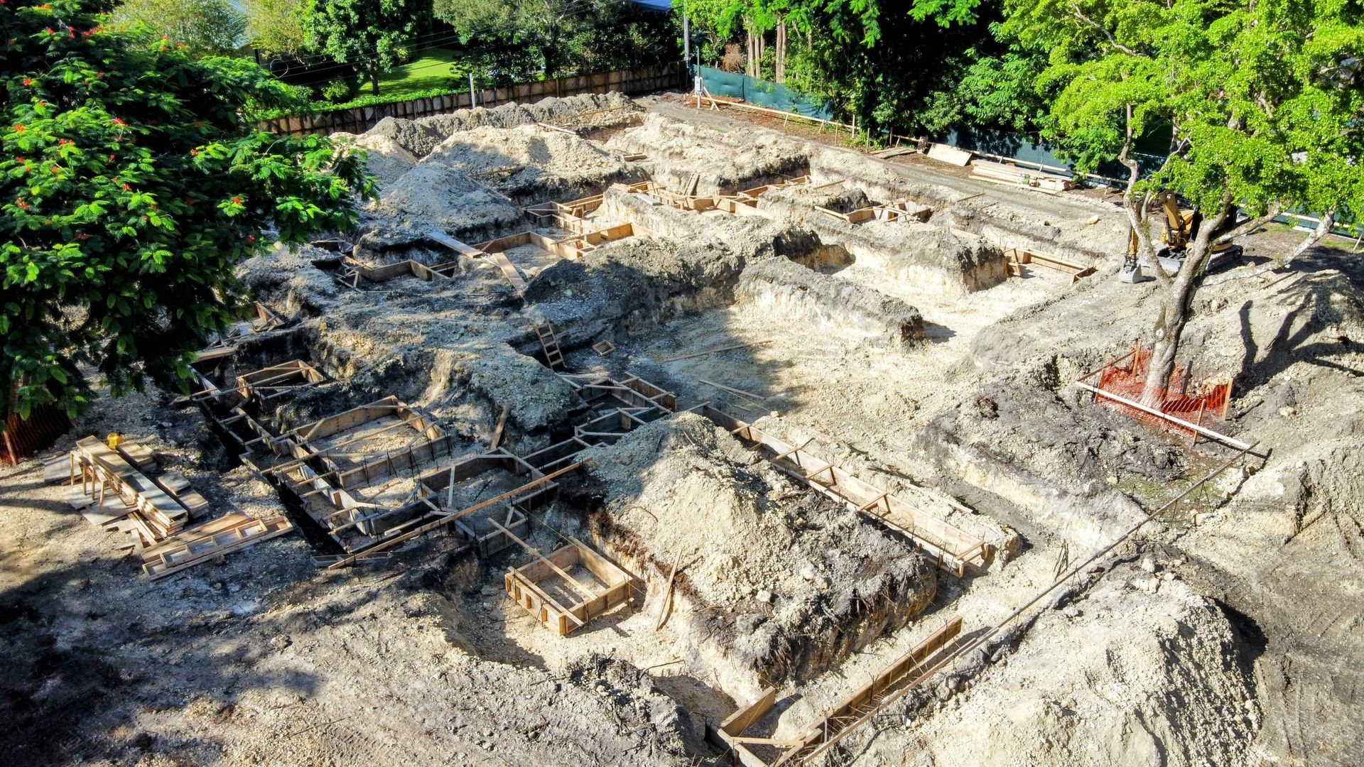 Archaeological excavation site, dirt, rectangular foundations, debris, surrounded by trees.