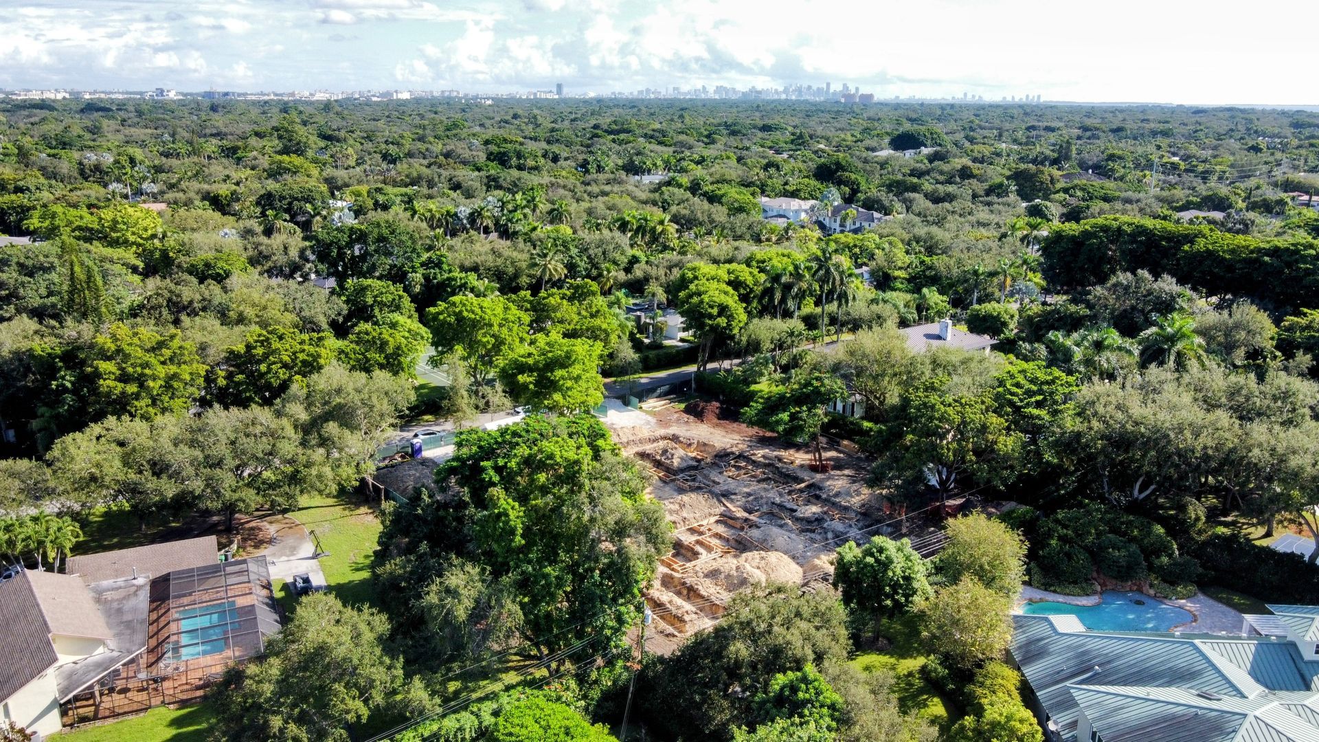 Aerial view of a cleared construction site surrounded by trees and houses, with a city skyline in the distance.