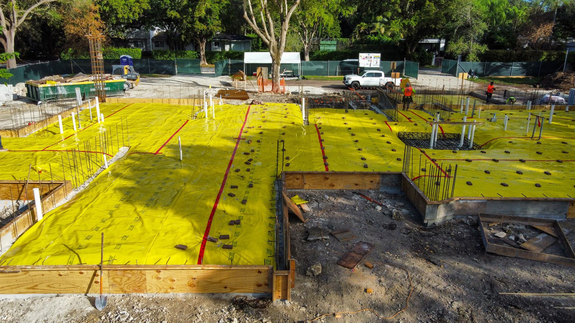 Construction site with yellow ground cover, wooden frames, and pipes. Workers and a truck are visible in the background.