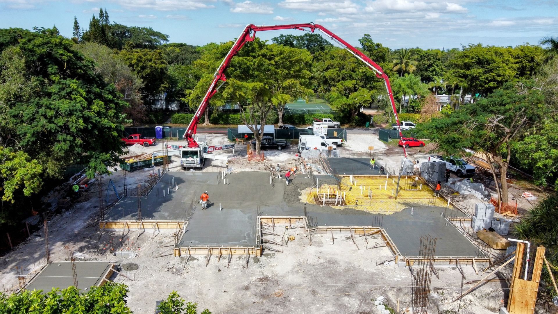 Construction site with concrete being poured, red pump truck, workers, green trees.