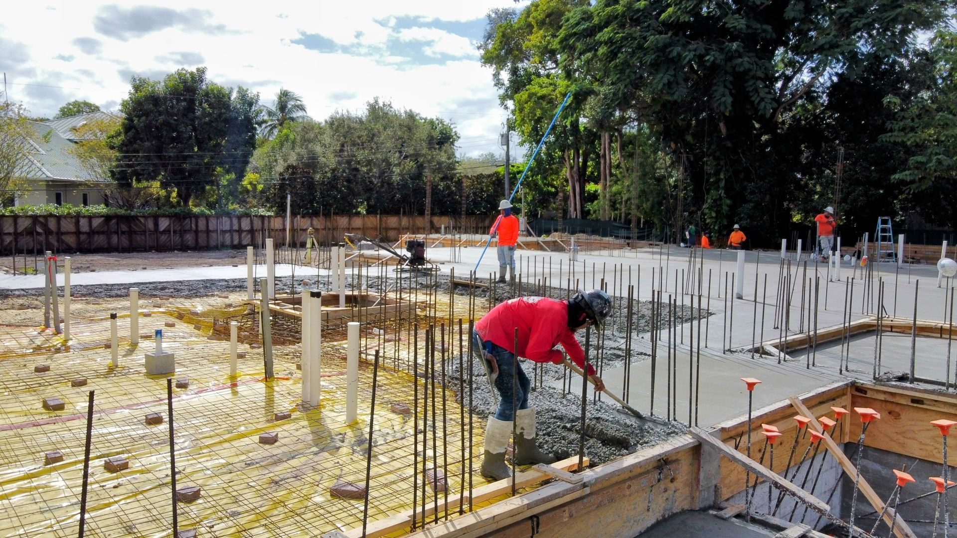 Construction workers pouring concrete foundation at a building site; includes rebar, wooden forms, and pipes.
