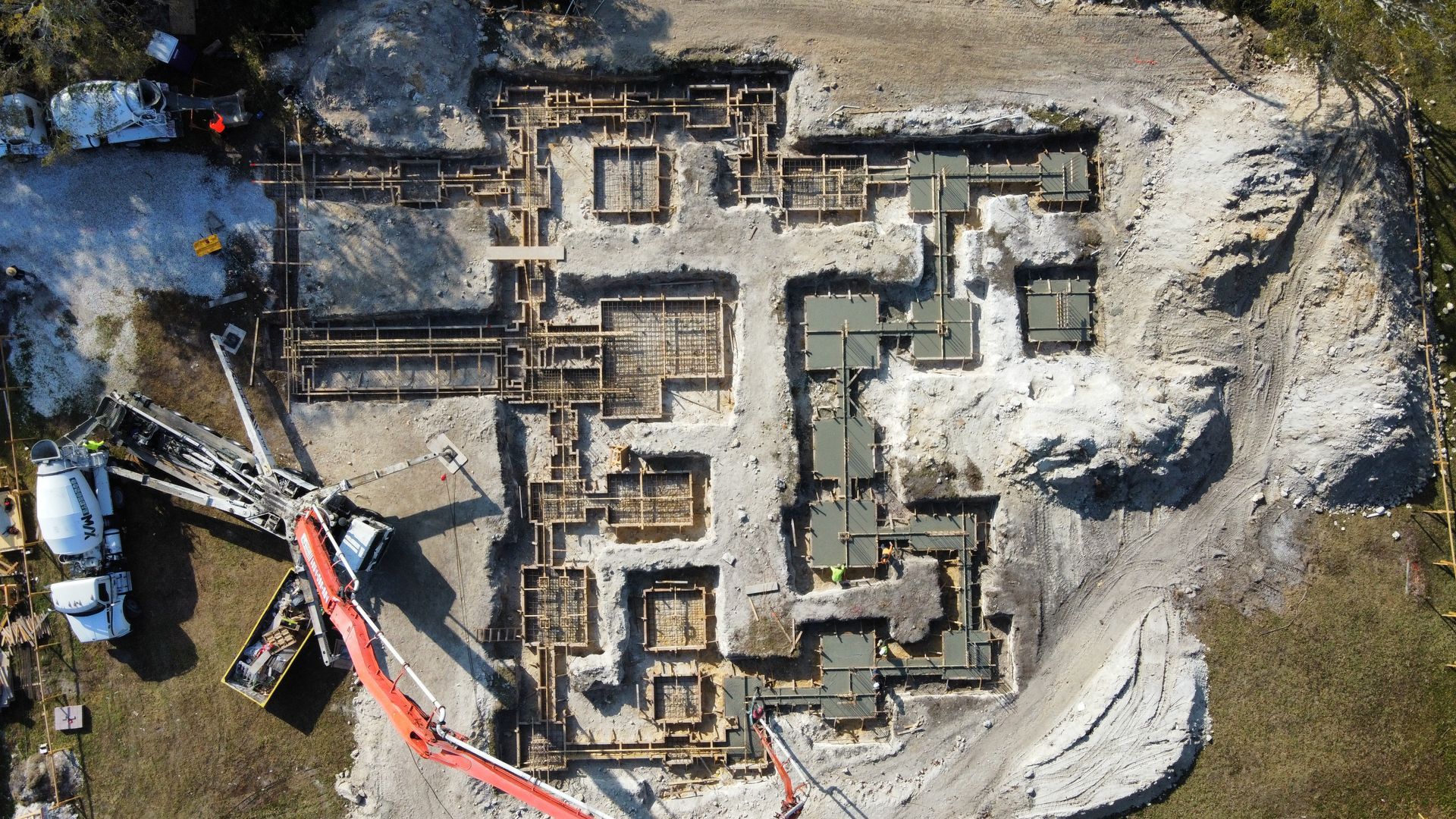 Overhead view of a construction site with excavated foundations and concrete forms. A cement truck pours concrete.